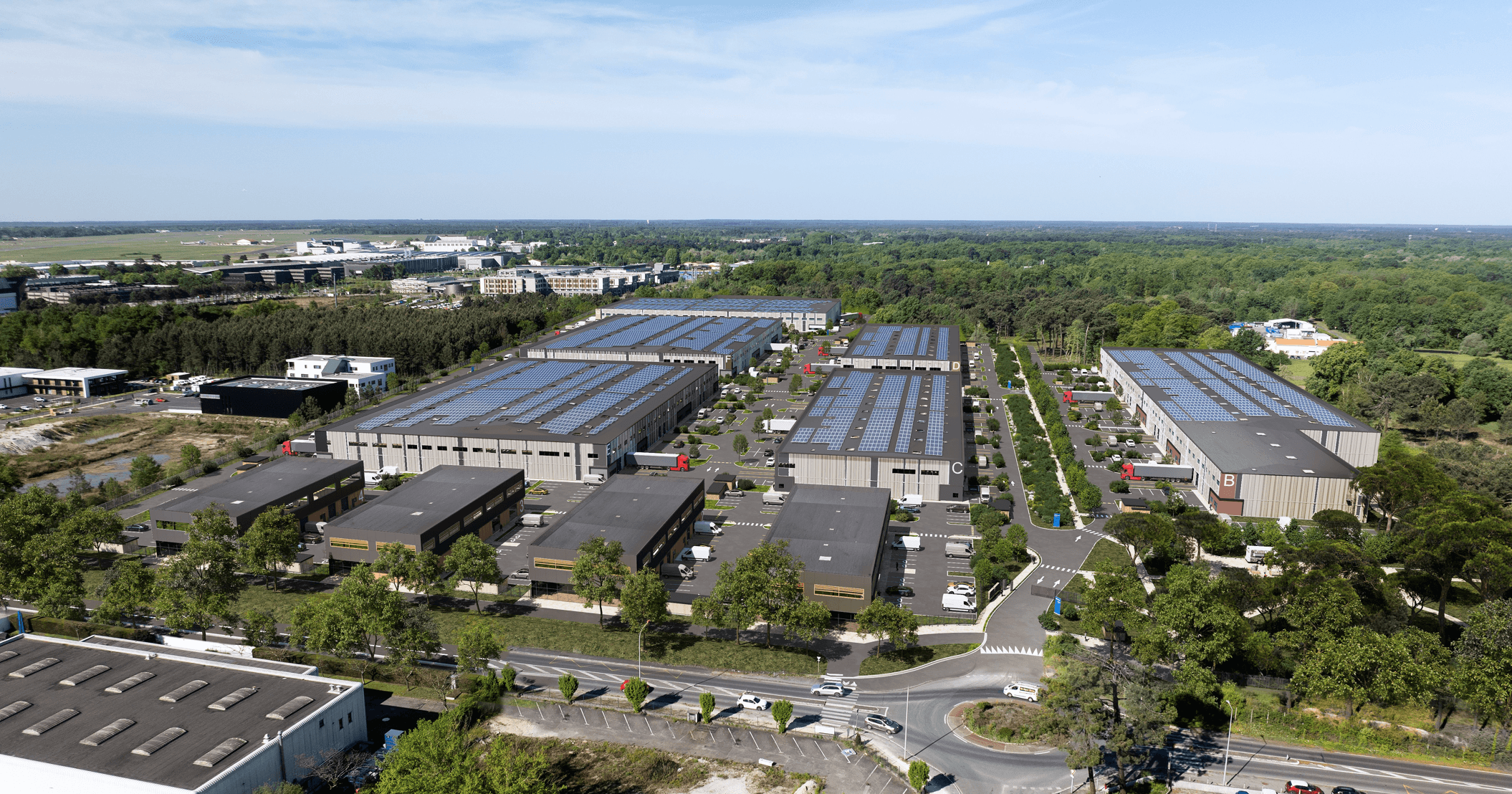 Aerial view of a large industrial complex with multiple warehouse buildings, solar panels on roofs, surrounding trees, and cars parked along internal roads.