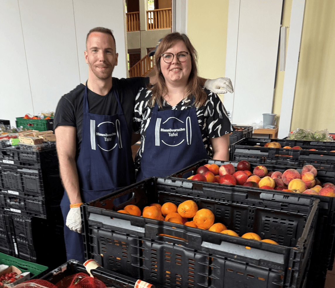 Two people wearing aprons and gloves stand by crates filled with fruit at an indoor food distribution or market setting.