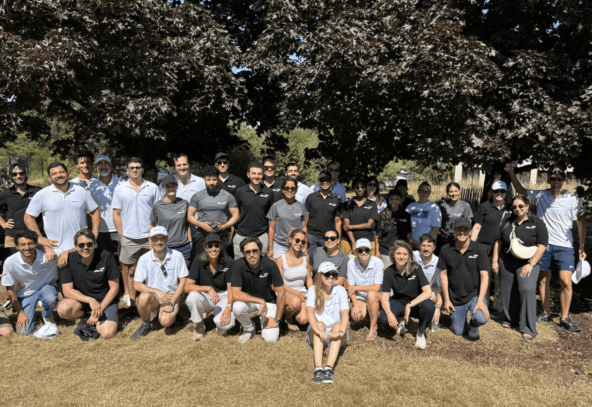 A group of people wearing black and white shirts pose together outdoors on dry grass in front of leafy trees, under sunny weather.