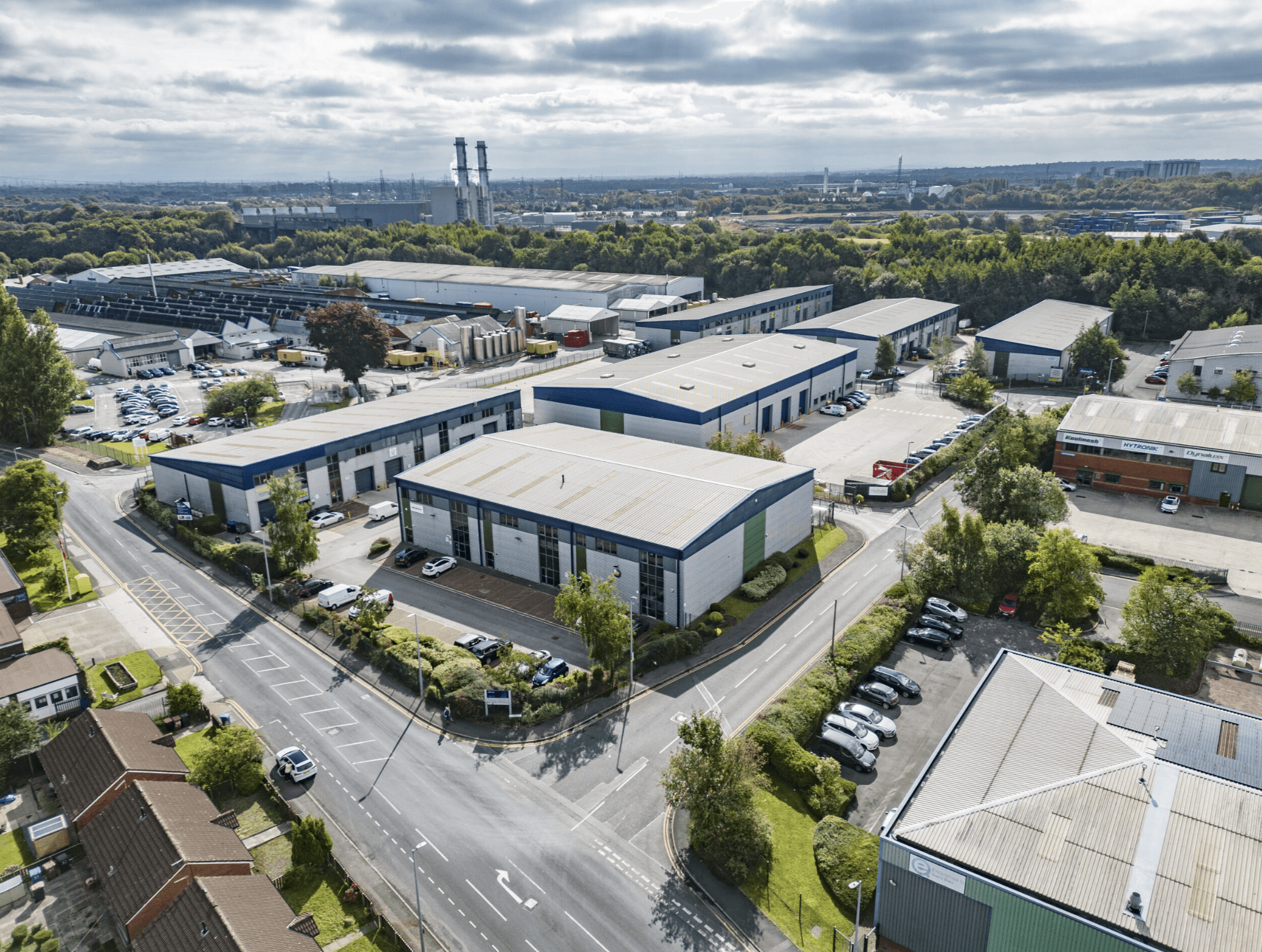 Aerial view of an industrial estate with multiple warehouses, surrounding greenery, parked cars, and a power plant in the background under a partly cloudy sky.