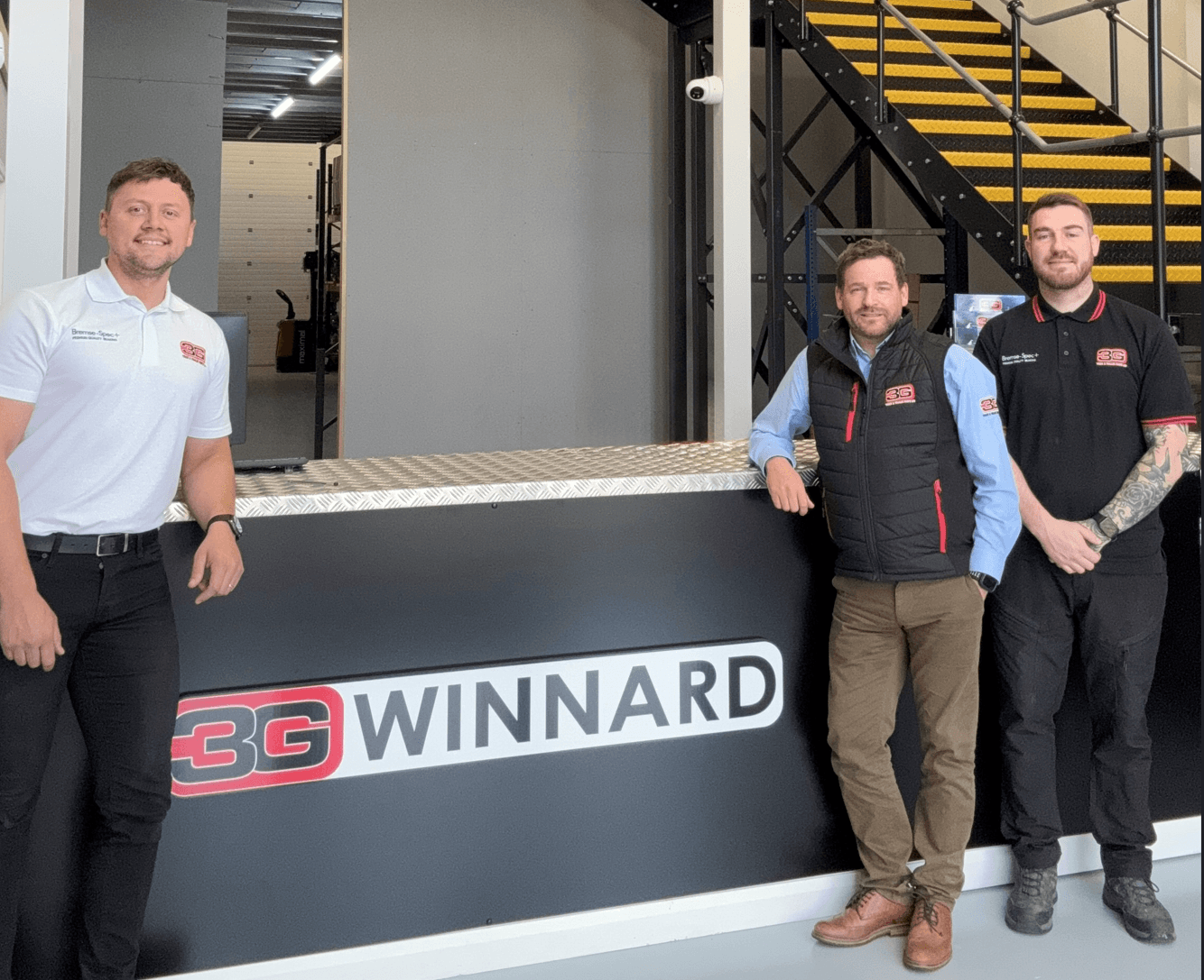 Three men stand in front of a reception desk with a 3G WINNARD sign, with stairs and an industrial interior in the background.