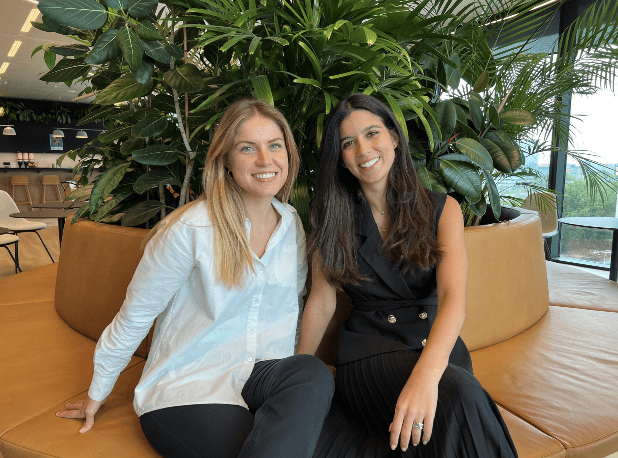 Two women smiling and sitting on a tan bench in front of large green plants in a modern indoor setting.
