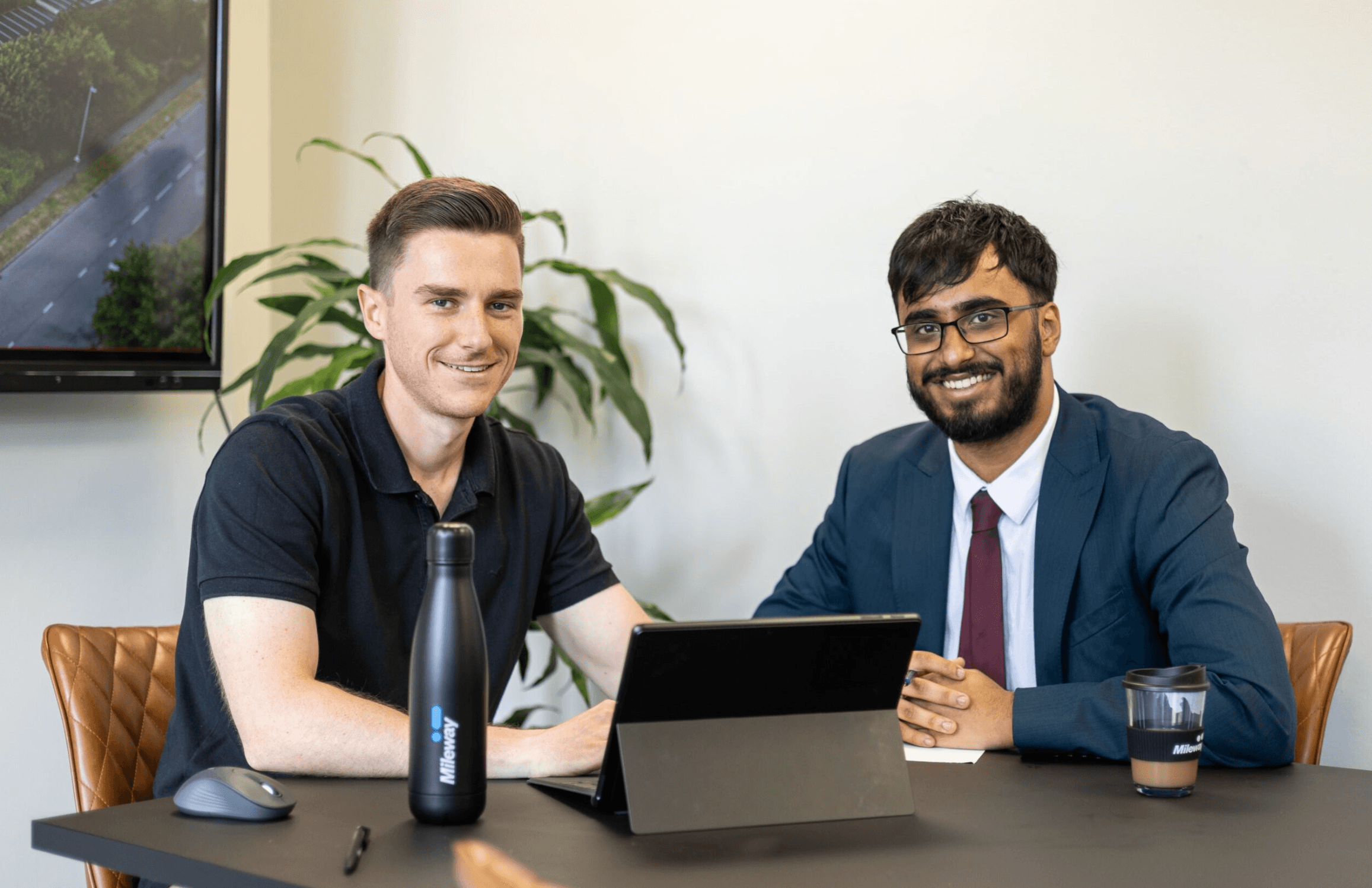 Two men sitting at a table with a laptop, water bottle, and coffee cup, smiling at the camera in a modern office setting.