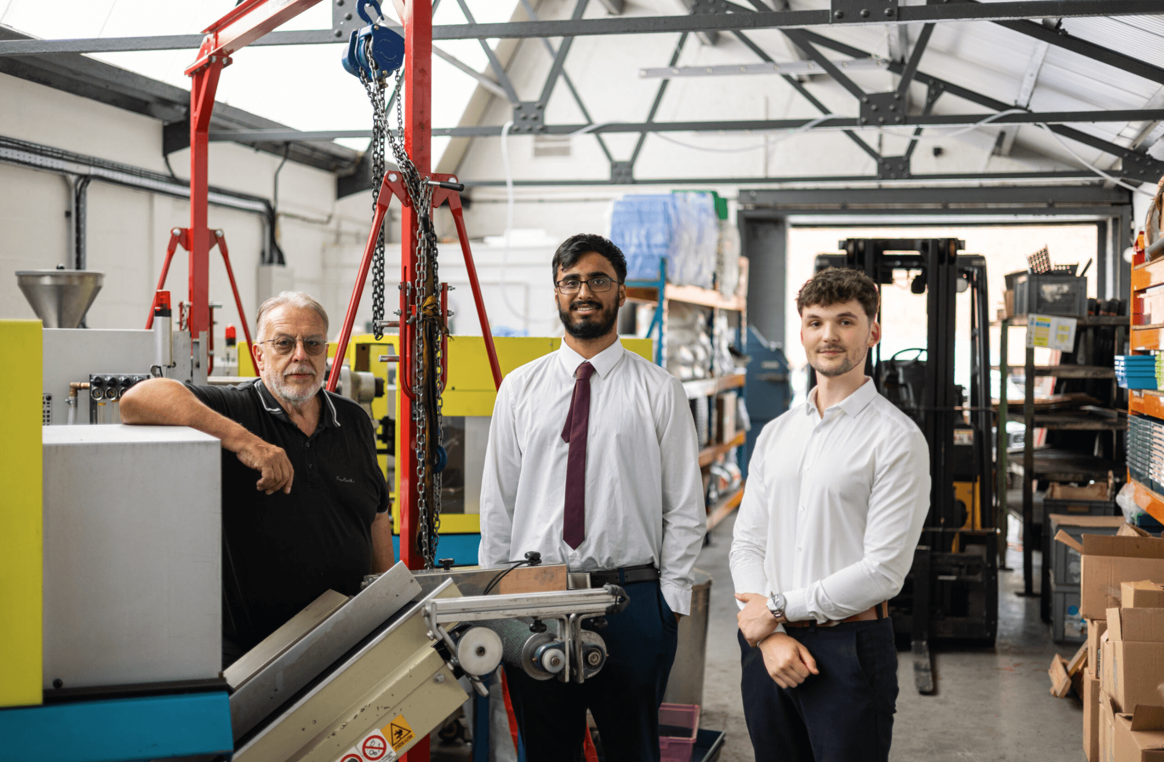 Three men stand in a manufacturing workshop next to machinery and shelving; two are in shirts and ties, one is in a black shirt.