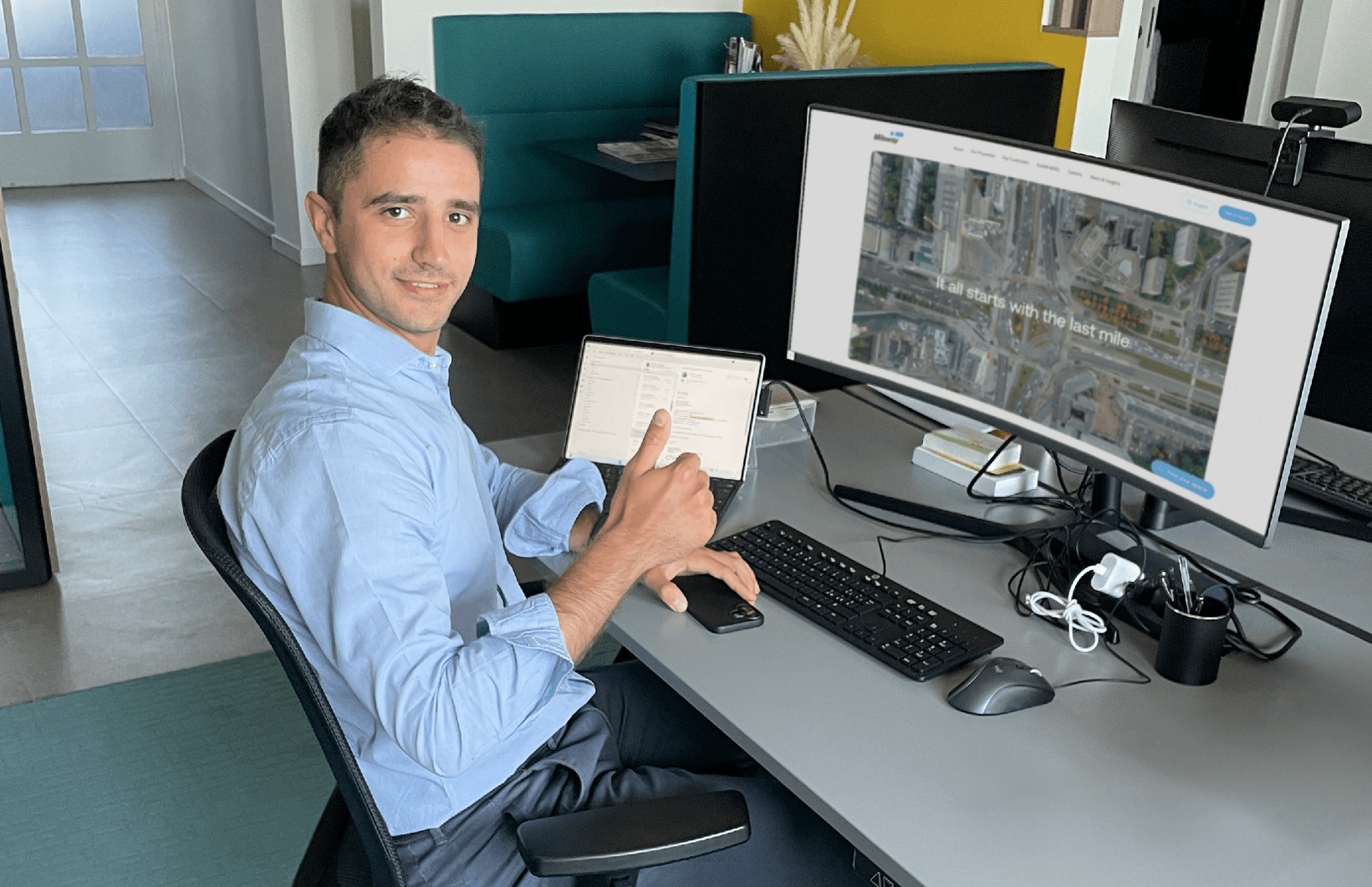 Man sitter vid ett skrivbord på ett kontor och gör tummen upp medan han arbetar på en dator med bildskärm och surfplatta.