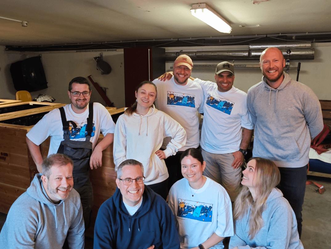 En grupp på nio vuxna, några i matchande t-shirts, poserar och ler inomhus framför en träkonstruktion.