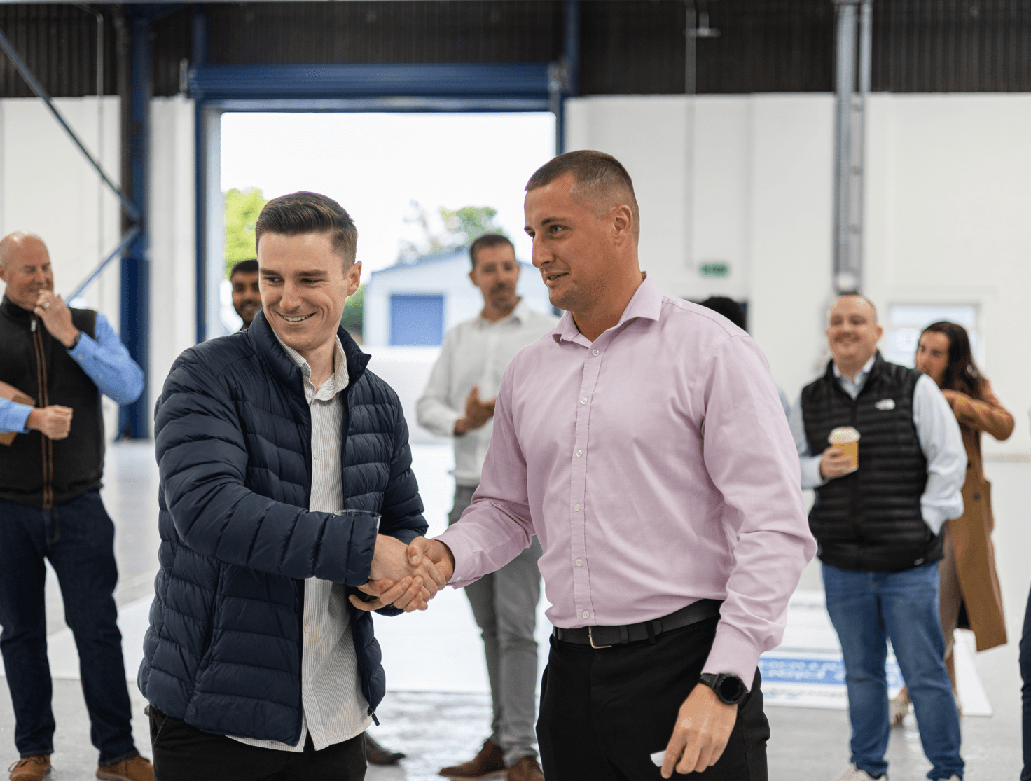 Två män skakar hand i förgrunden av en inomhusmiljö, medan flera personer står i bakgrunden och tittar på och ler.