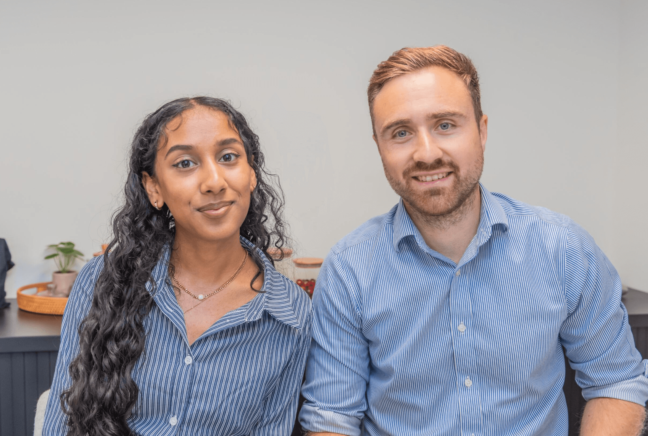 Två personer sitter sida vid sida, båda bär blå skjortor med knappar, tittar in i kameran och ler i en kontorsmiljö.