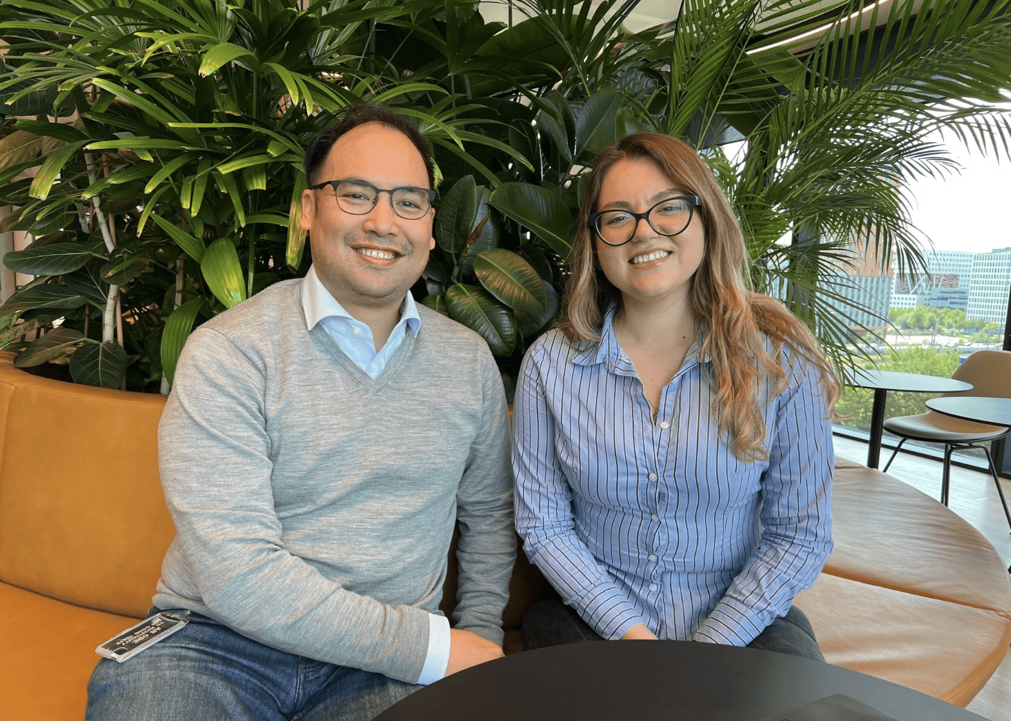 Två personer sitter i en brun soffa framför gröna växter, båda bär glasögon och ler mot kameran i en modern inomhusmiljö.