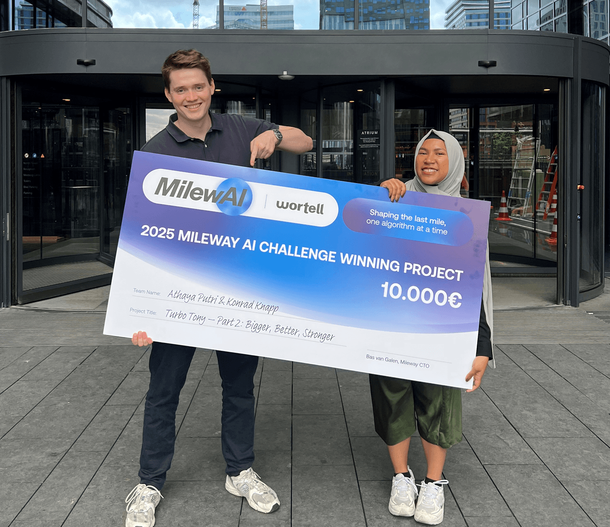 Två personer står utanför en byggnad och håller i en stor check på 10.000 euro som belöning för att ha vunnit 2025 års Mileway AI Challenge.
