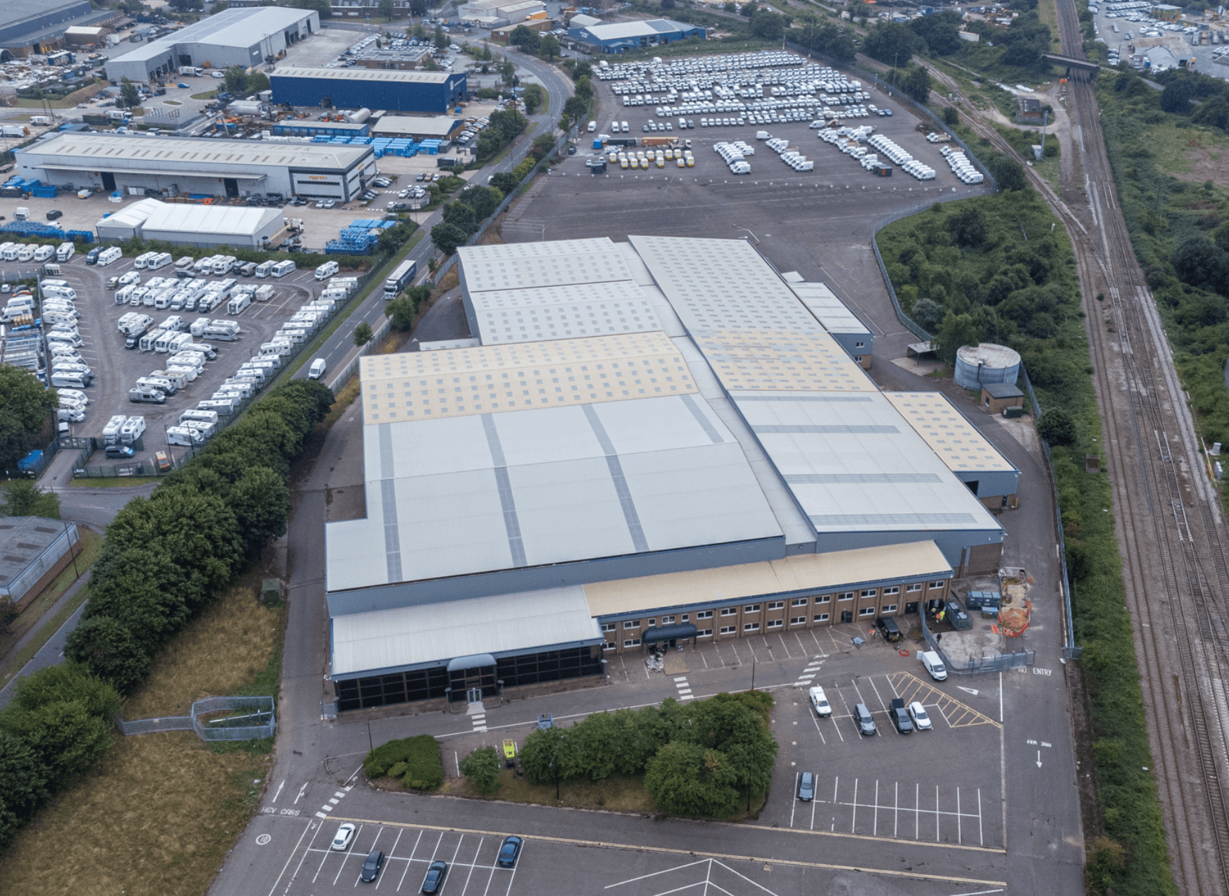 Flygbild över ett stort industrilager med en parkeringsplats framför, omgiven av parkerade fordon, träd och närliggande byggnader. Järnvägsspår löper längs bildens högra sida.