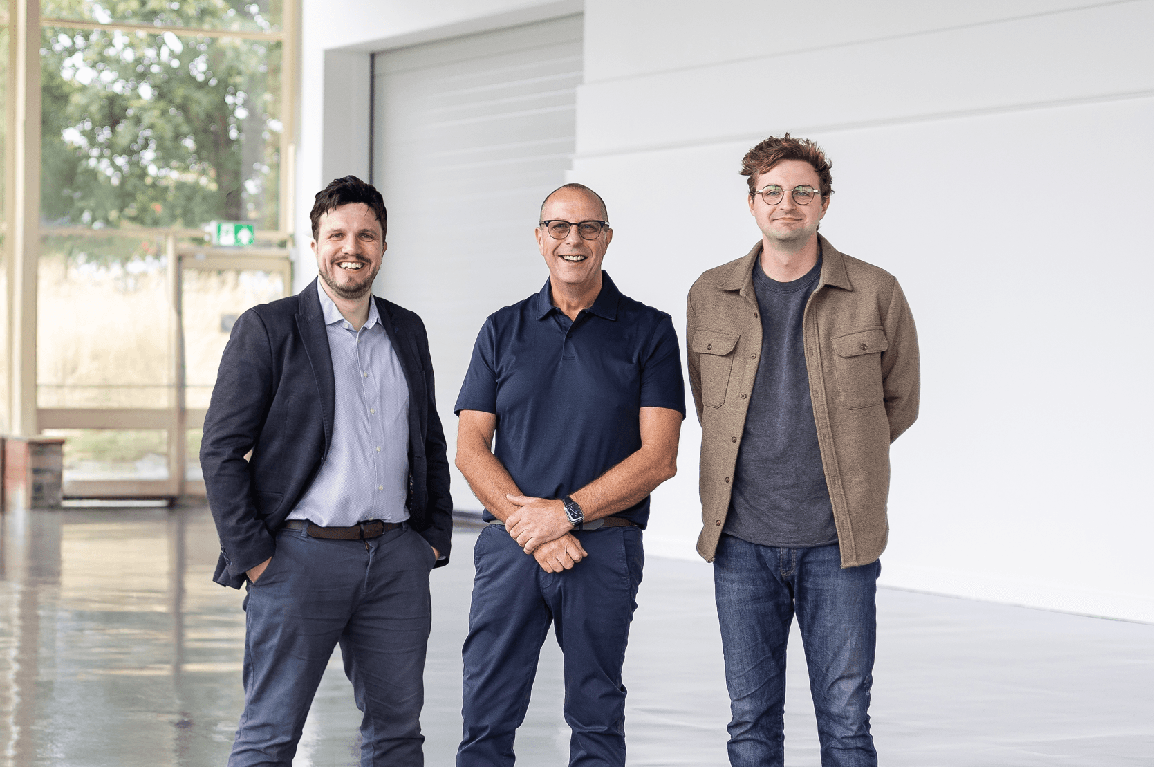 Tre män står sida vid sida inomhus i en ljus, modern lokal med stora fönster och vita väggar, alla vänder sig mot kameran och ler.