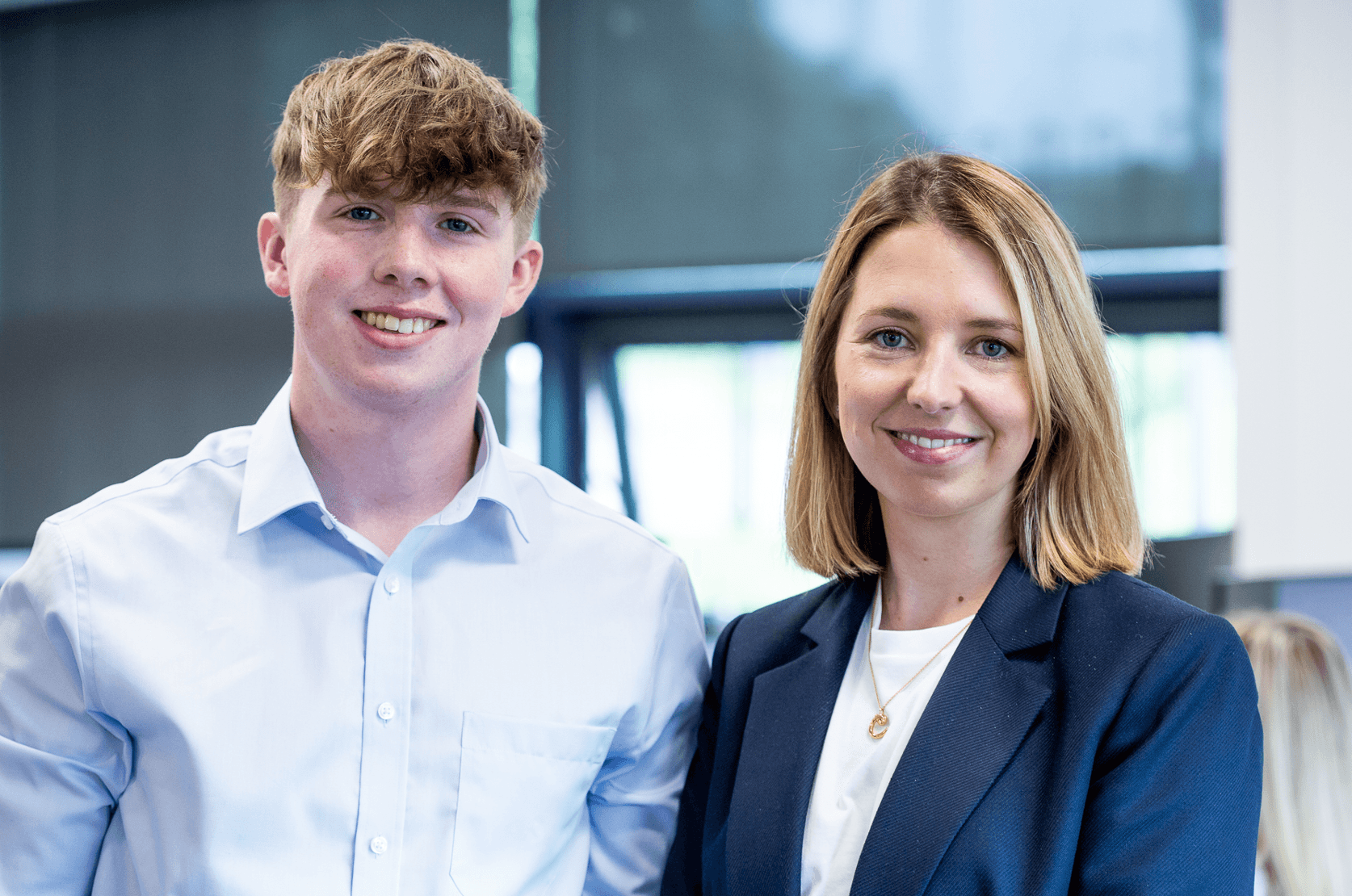 En ung man och en kvinna, båda klädda i affärskläder, står sida vid sida inomhus och ler mot kameran.