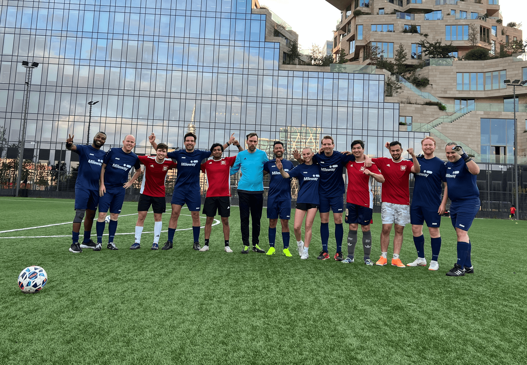 Un gruppo di giocatori di calcio con maglie blu e rosse sta a braccetto su un campo, sorridendo alla telecamera con un moderno edificio di vetro sullo sfondo.