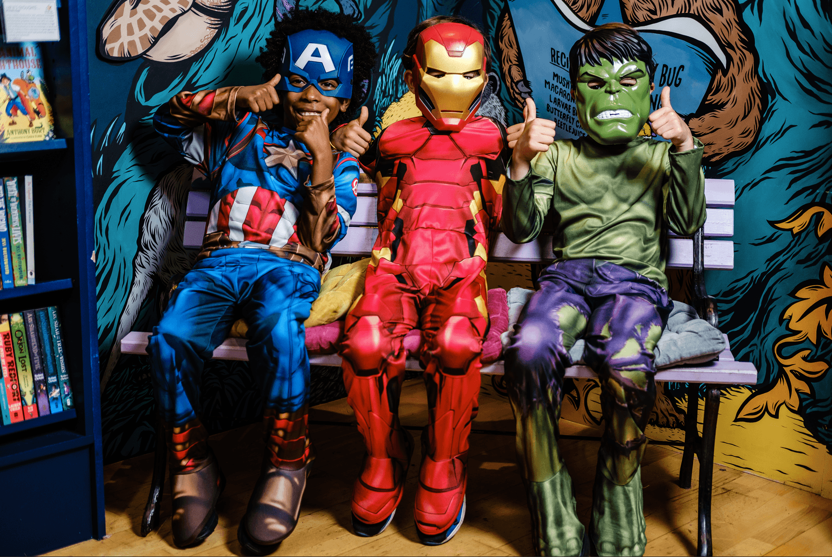 Three children in Captain America, Iron Man, and Hulk costumes sit on a bench, giving thumbs up, in front of a comic-themed mural.