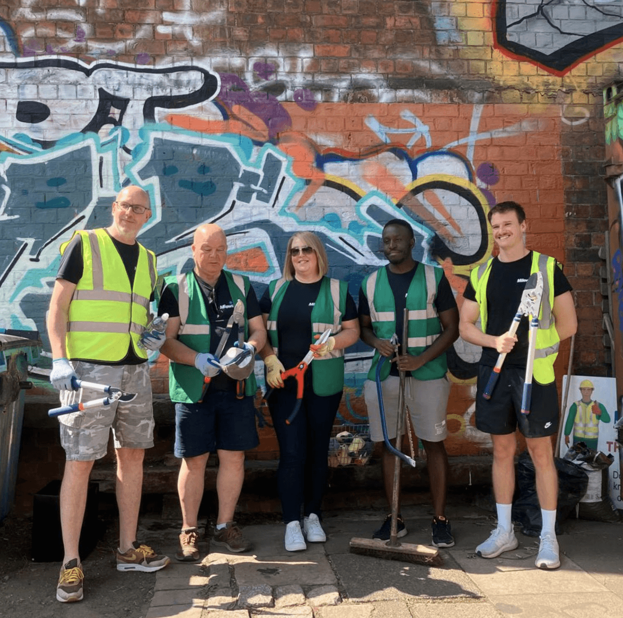 Five people wearing safety vests and gloves stand in front of a graffiti wall, holding litter-picking tools and a broom, posing for a group photo outdoors.
