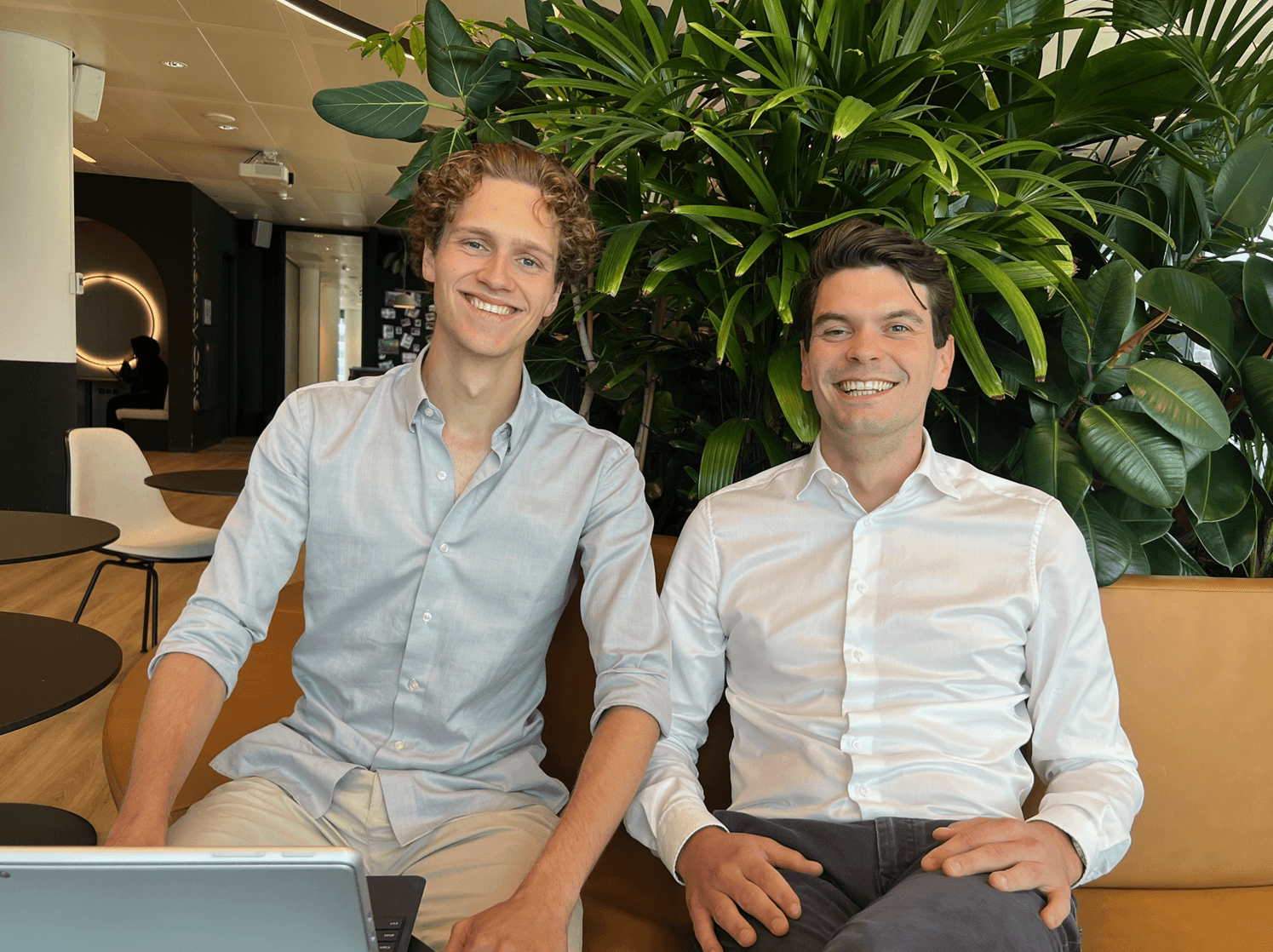 Two men sit on a tan couch in a modern office space with green plants behind them, both smiling, wearing light-colored shirts. A laptop is visible in the foreground.