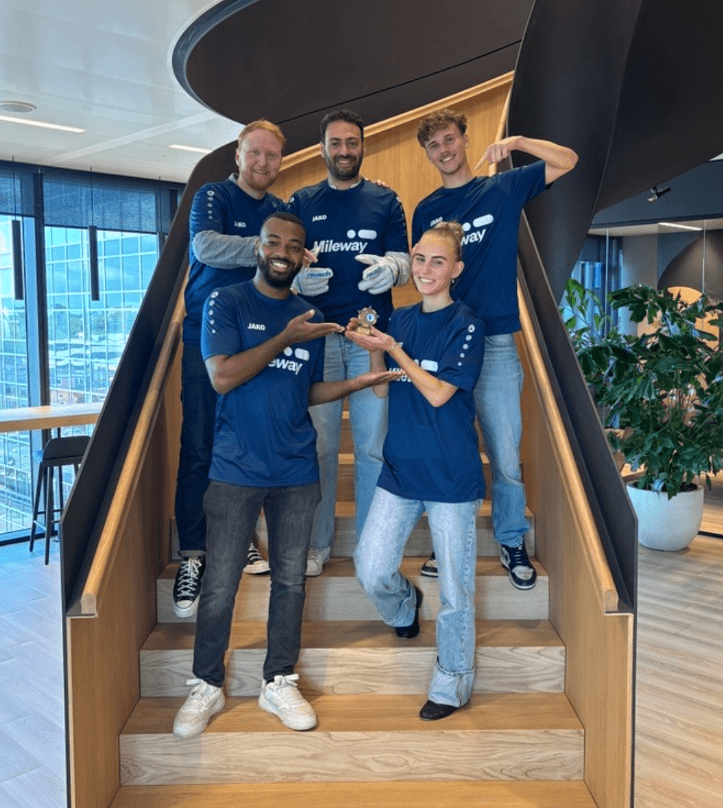 Five people wearing matching blue shirts stand on wooden stairs, smiling and gesturing towards a small trophy held by the person in front.