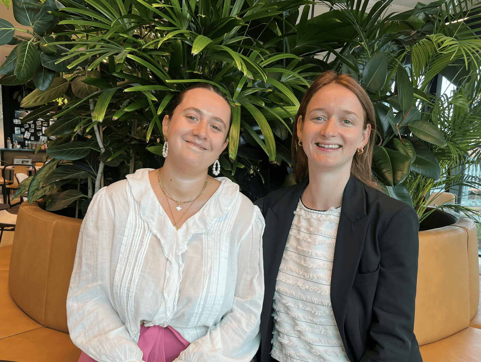 Two women sit side by side indoors in front of large green plants, both smiling at the camera. One wears a white blouse; the other wears a white top with a black jacket.