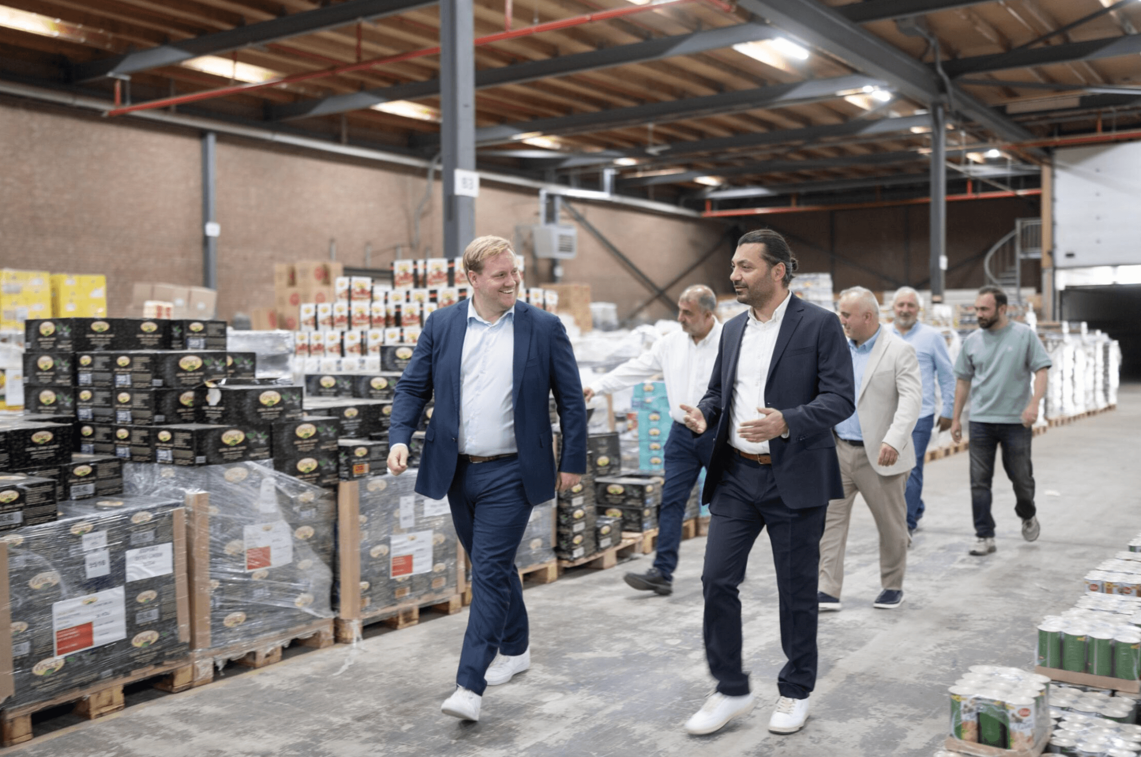 A group of men in business attire walk through a warehouse with pallets of packaged goods stacked around them.