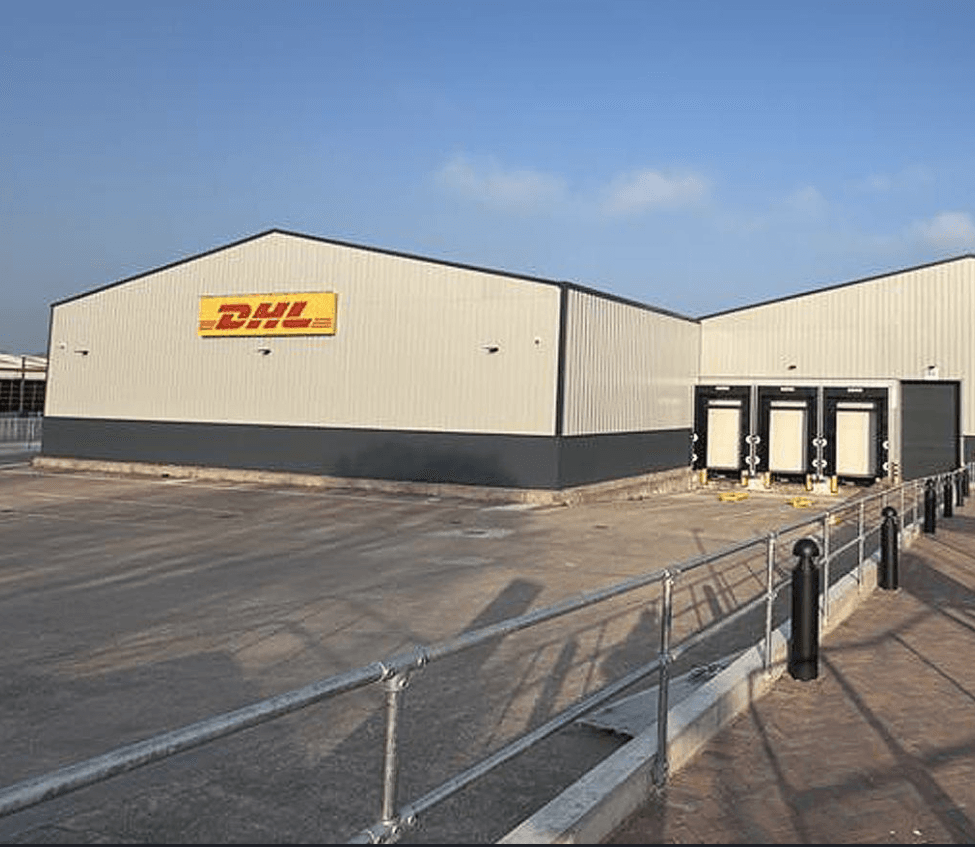 A DHL warehouse building with a yellow DHL sign, three loading docks, and an empty paved lot in front under a clear sky.