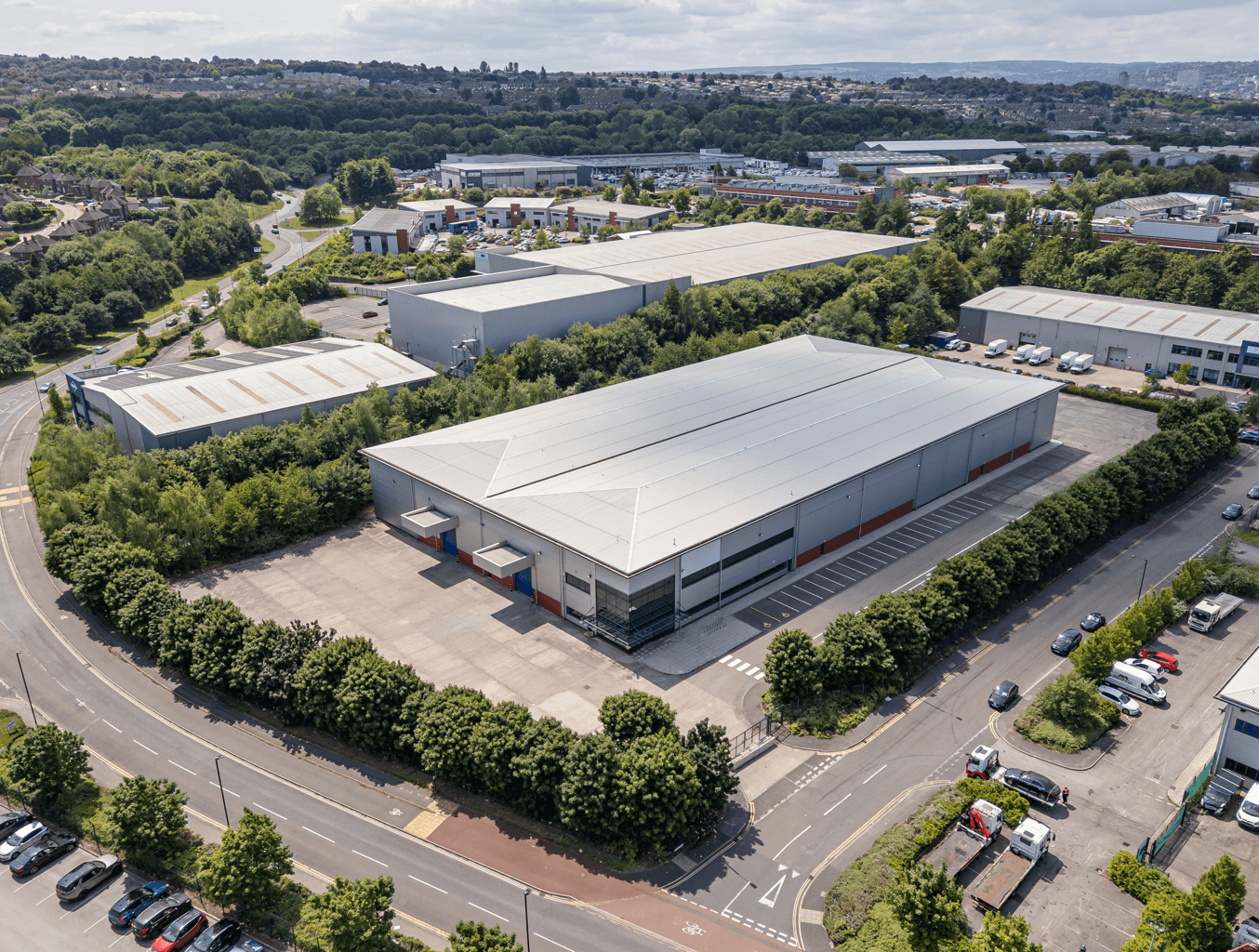 Aerial view of a large industrial warehouse with loading docks, surrounded by trees, parking lots, and adjacent roads in a business park area.