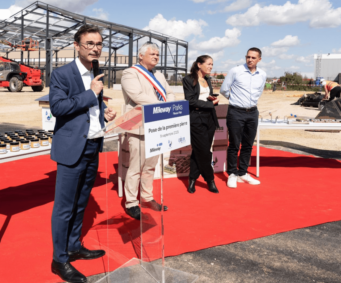Four people stand on a red carpet at an outdoor event with a construction site in the background. One man speaks at a podium labeled “Mileway Parks.”.
