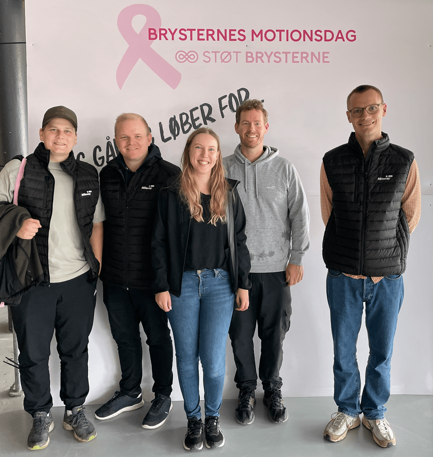 Five people stand in front of a banner for Brysternes Motionsdag, an event supporting breast health awareness. The group is casually dressed and smiling at the camera.