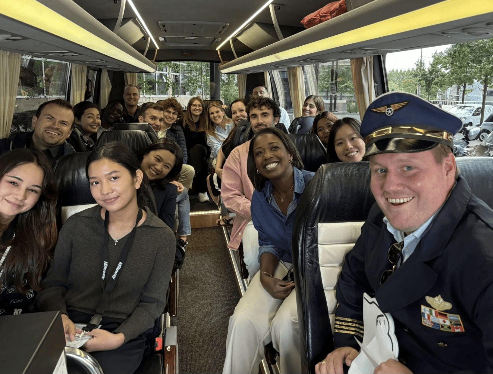 A group of people, including a man dressed as a pilot, sit and smile at the camera inside a bus with large windows.