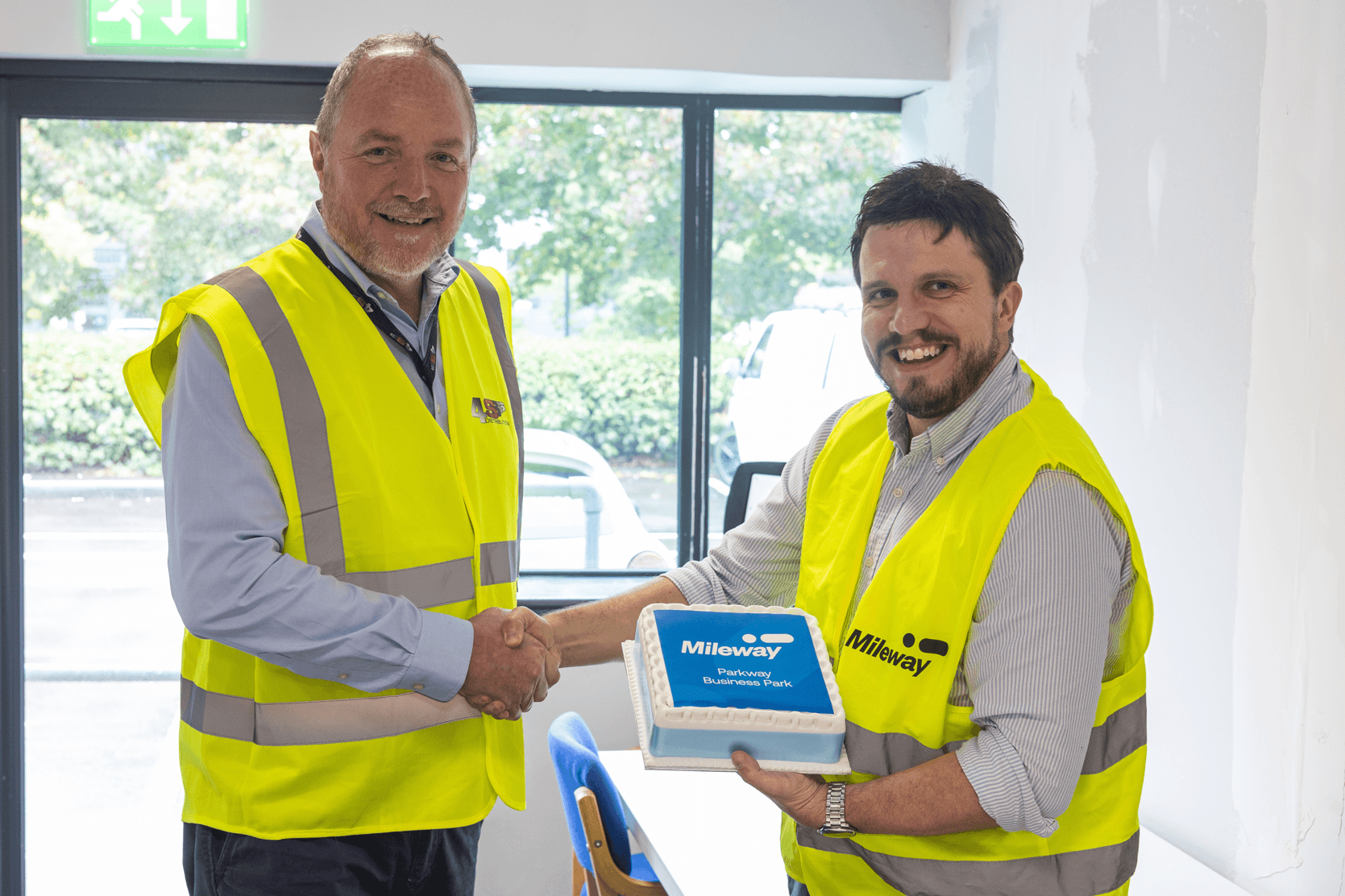 Two men wearing yellow high-visibility vests shake hands while one holds a rectangular blue cake with Mileway written on it, standing indoors near a window.