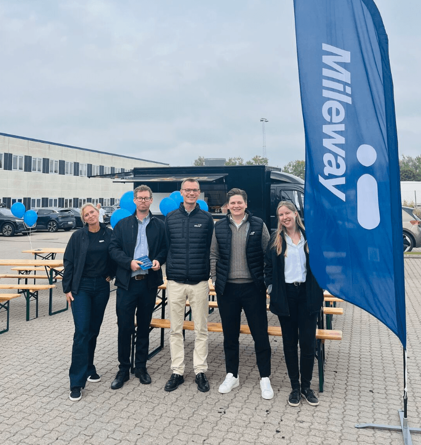 Five people stand in front of a black van and picnic tables with blue balloons. A blue flag with the Mileway logo is displayed on the right. Industrial building in background.