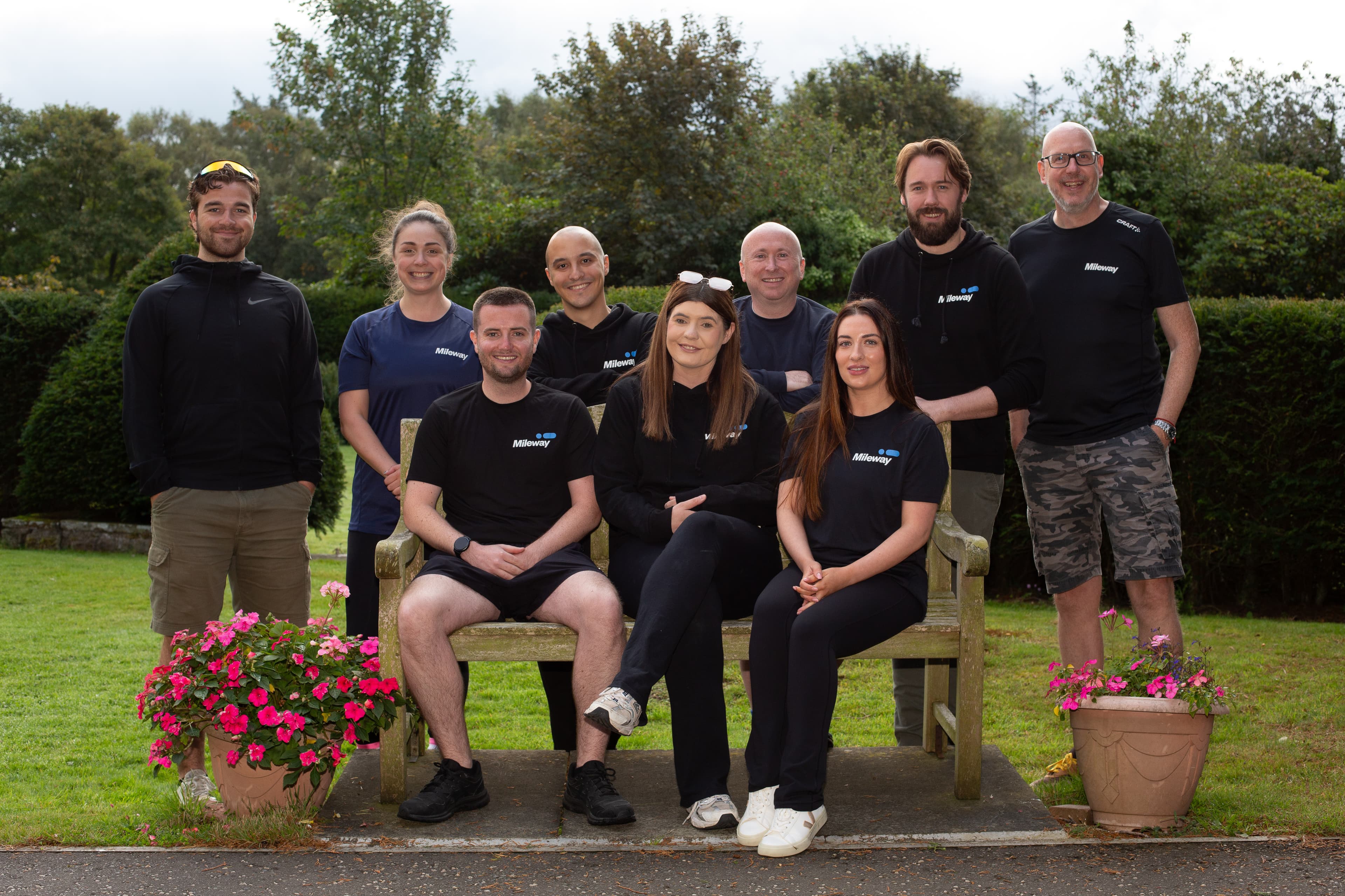 A group of nine adults, wearing black shirts, poses together outside on and around a wooden bench, with green foliage and flower pots in the background.