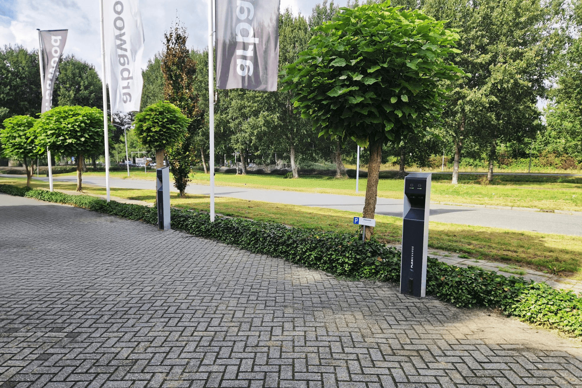 Two electric vehicle charging stations stand on a paved area beside a hedge, with trees, flags, and a road in the background.