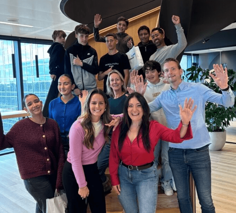 A group of thirteen people, some standing and some sitting on stairs, are smiling and waving inside a modern office space with large windows.