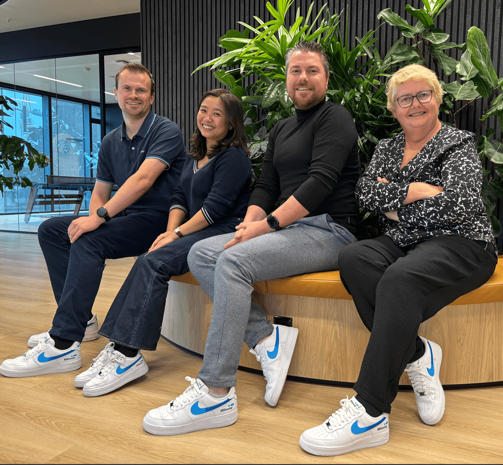 Four people sit side by side on a curved wooden bench indoors, smiling. Three wear matching white sneakers with blue swooshes. There are green plants behind them.