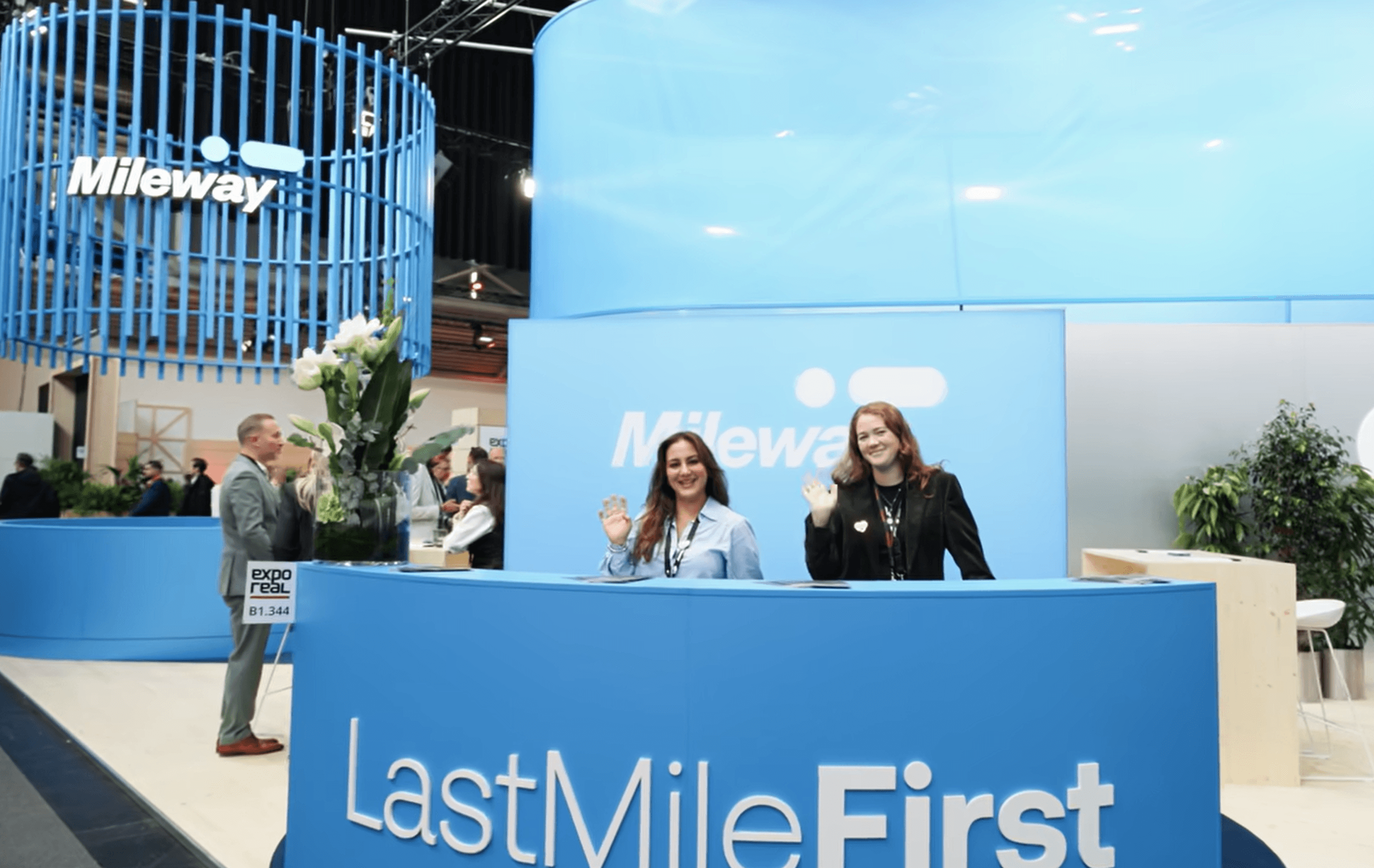 Two women stand and wave at a blue reception desk with LastMileFirst and Mileway branding at an indoor exhibition or trade fair.