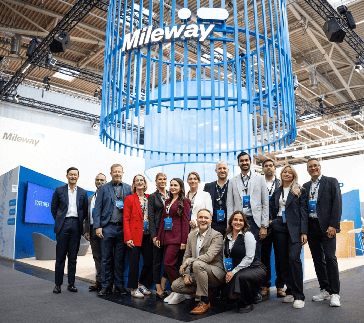 A group of people posing and smiling in front of a blue and white Mileway stand at a trade show or conference.