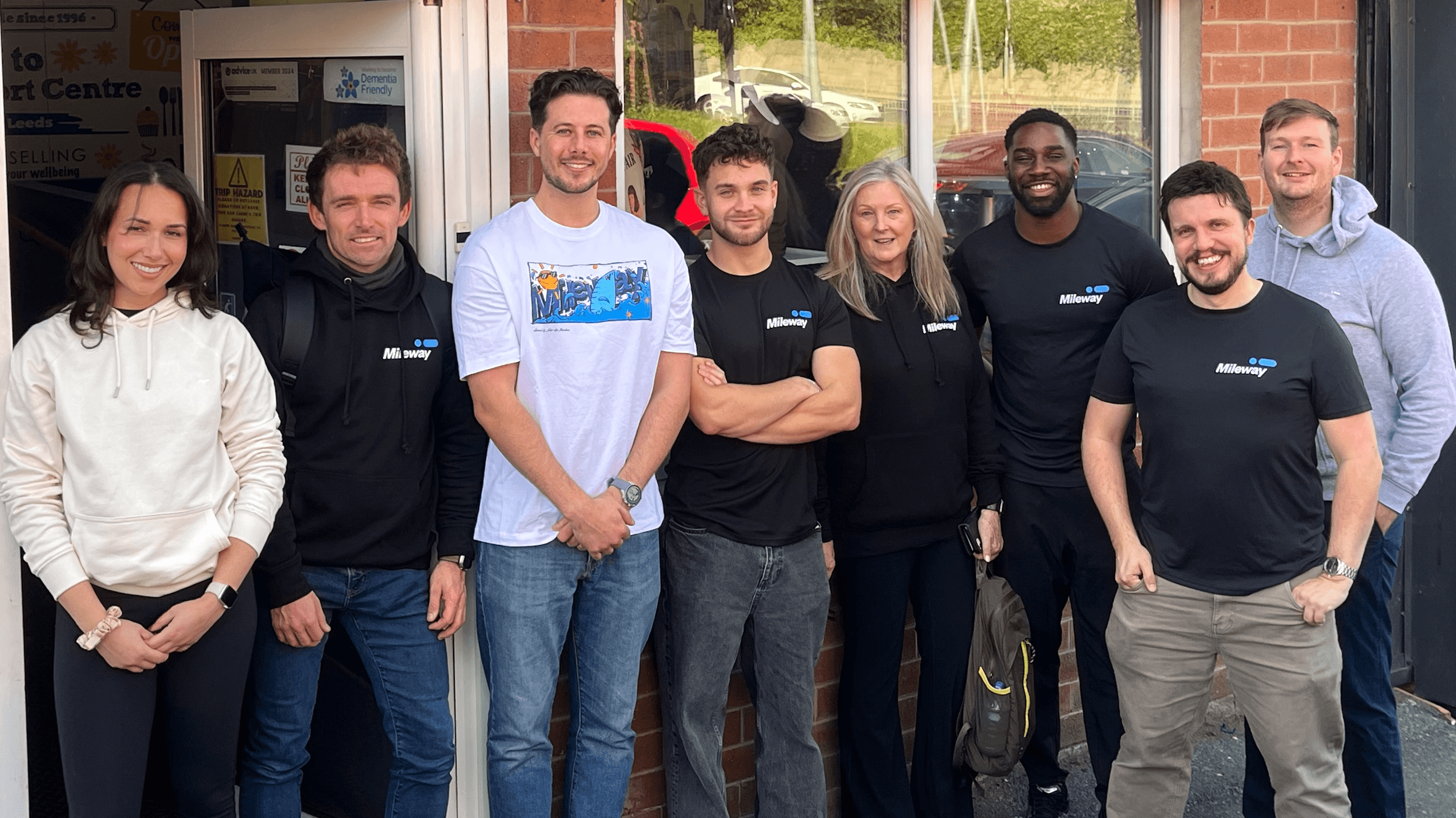 Eight adults stand side by side outside a brick building, posing for a group photo. Most are wearing black “AllMoney” shirts, and all are smiling at the camera.