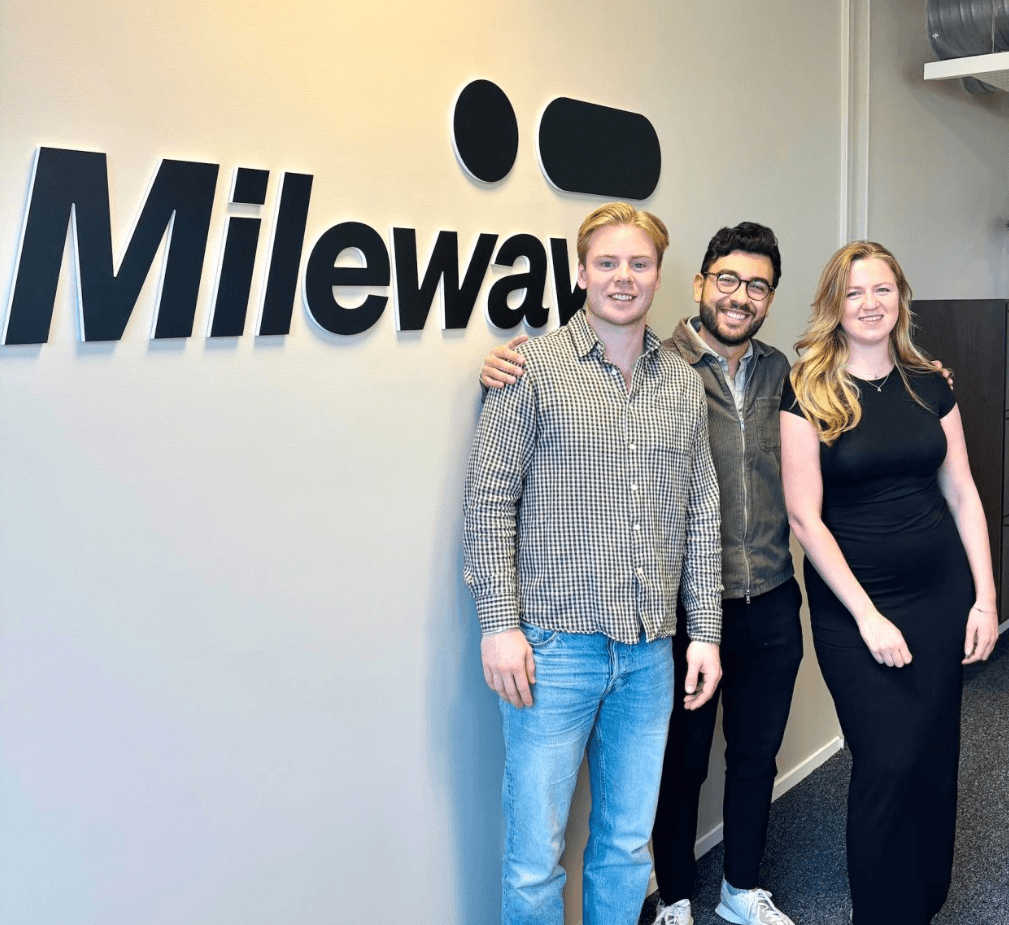 Three people stand smiling in front of a wall with the Mileway logo. The group includes two men and one woman, all dressed in casual to business-casual attire.