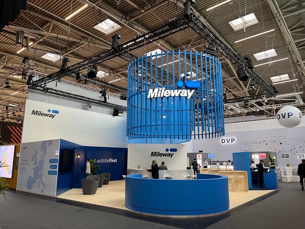 Mileway exhibition stand with blue and white design, circular reception desk, seating area, digital displays, and overhead signage in a large event hall.