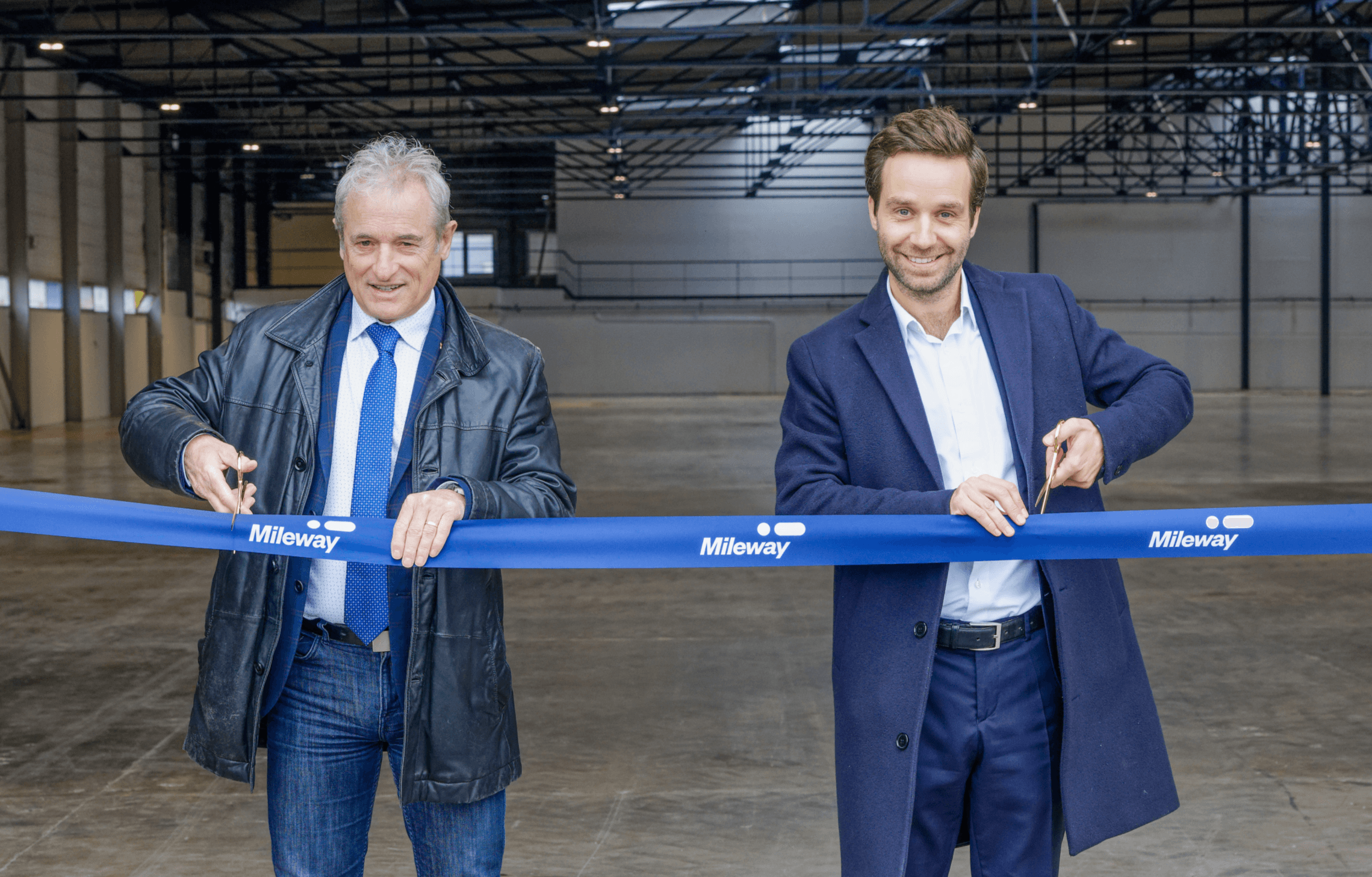 Two men in business attire cut a blue ribbon labelled Mileway in an empty industrial warehouse, marking an opening ceremony.