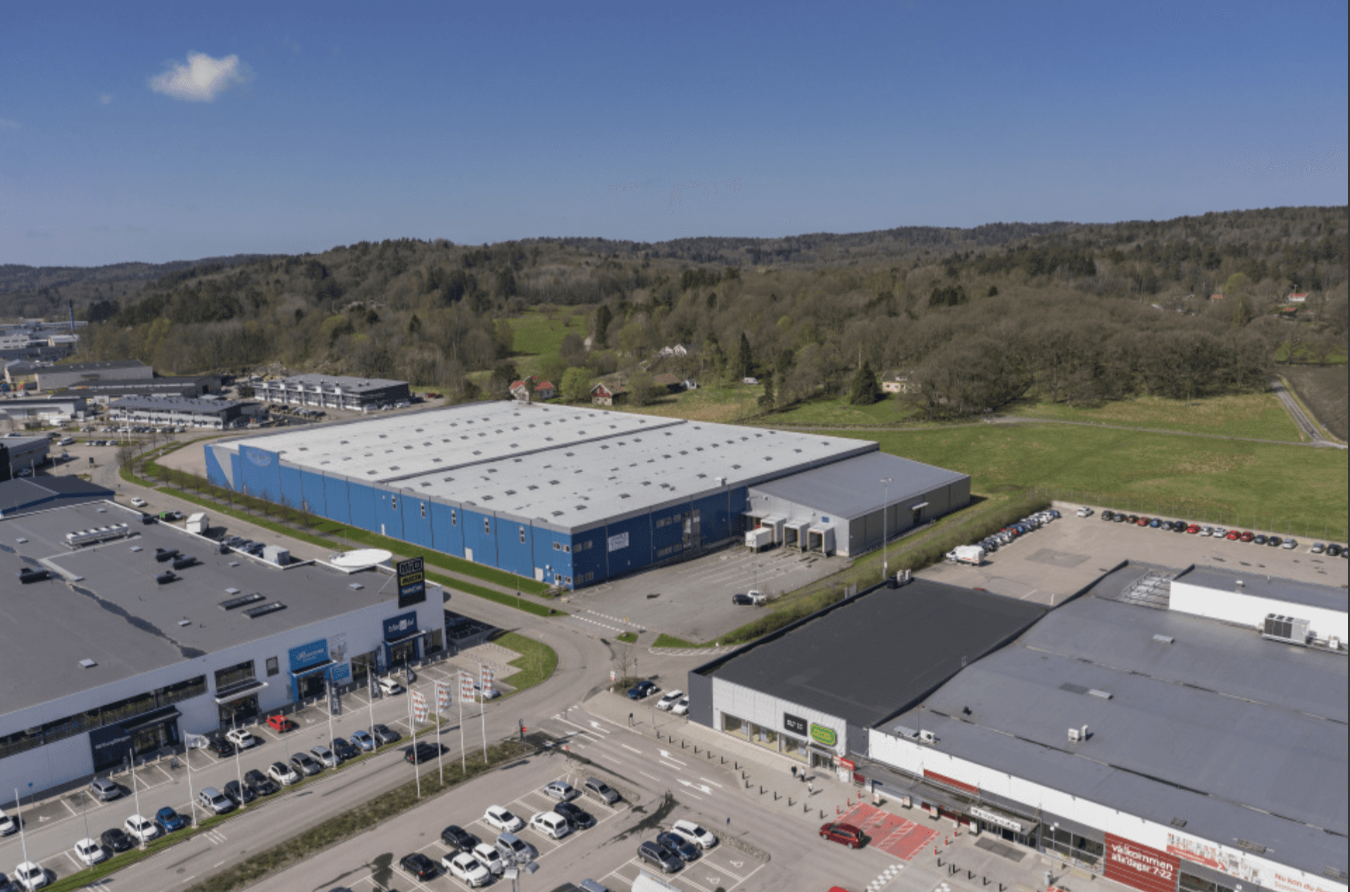 Aerial view of a commercial and industrial area with large warehouses, car parks, and nearby green fields under a clear blue sky.