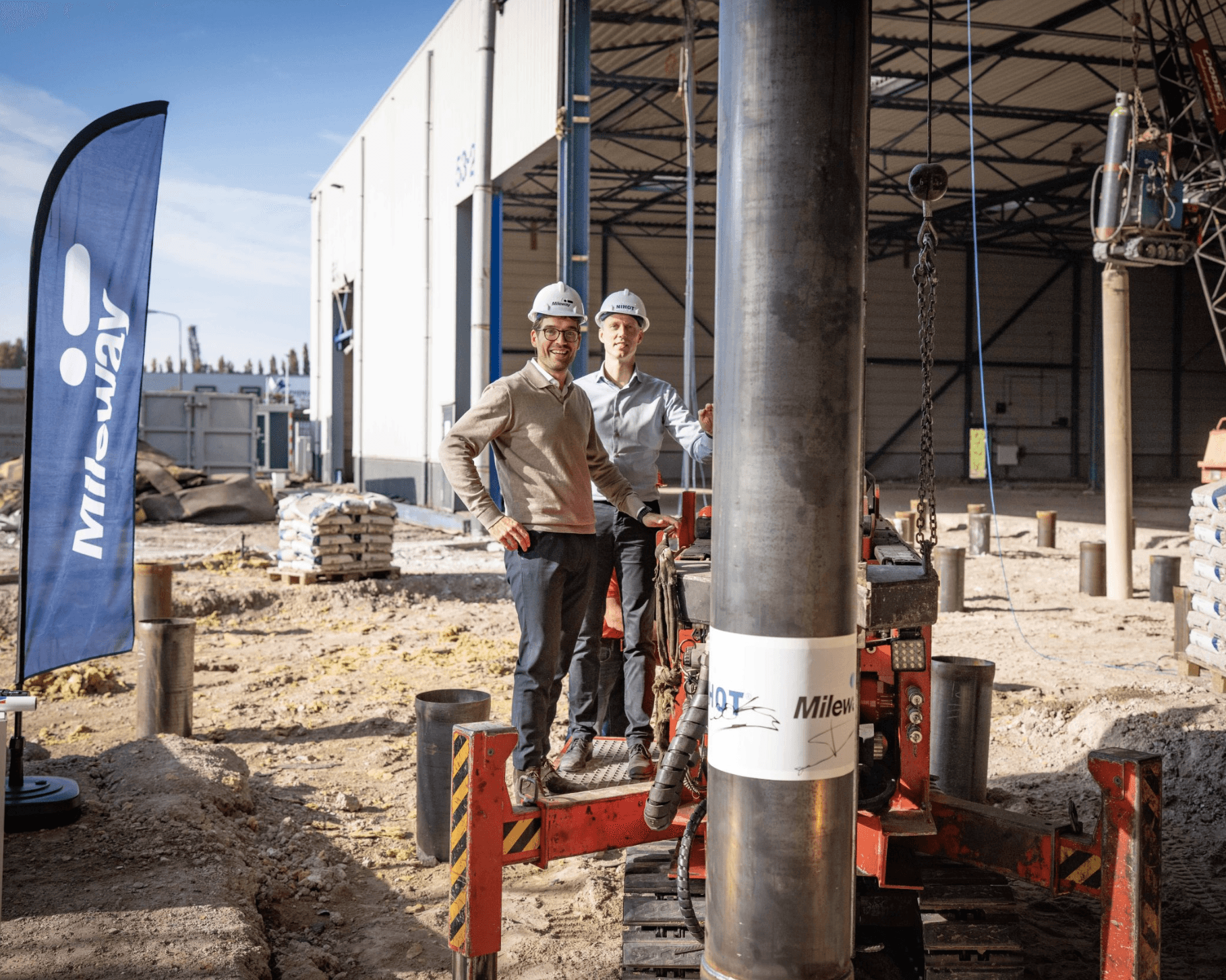 Two men in safety helmets stand next to industrial equipment at a construction site with a Mileway flag and warehouse in the background.