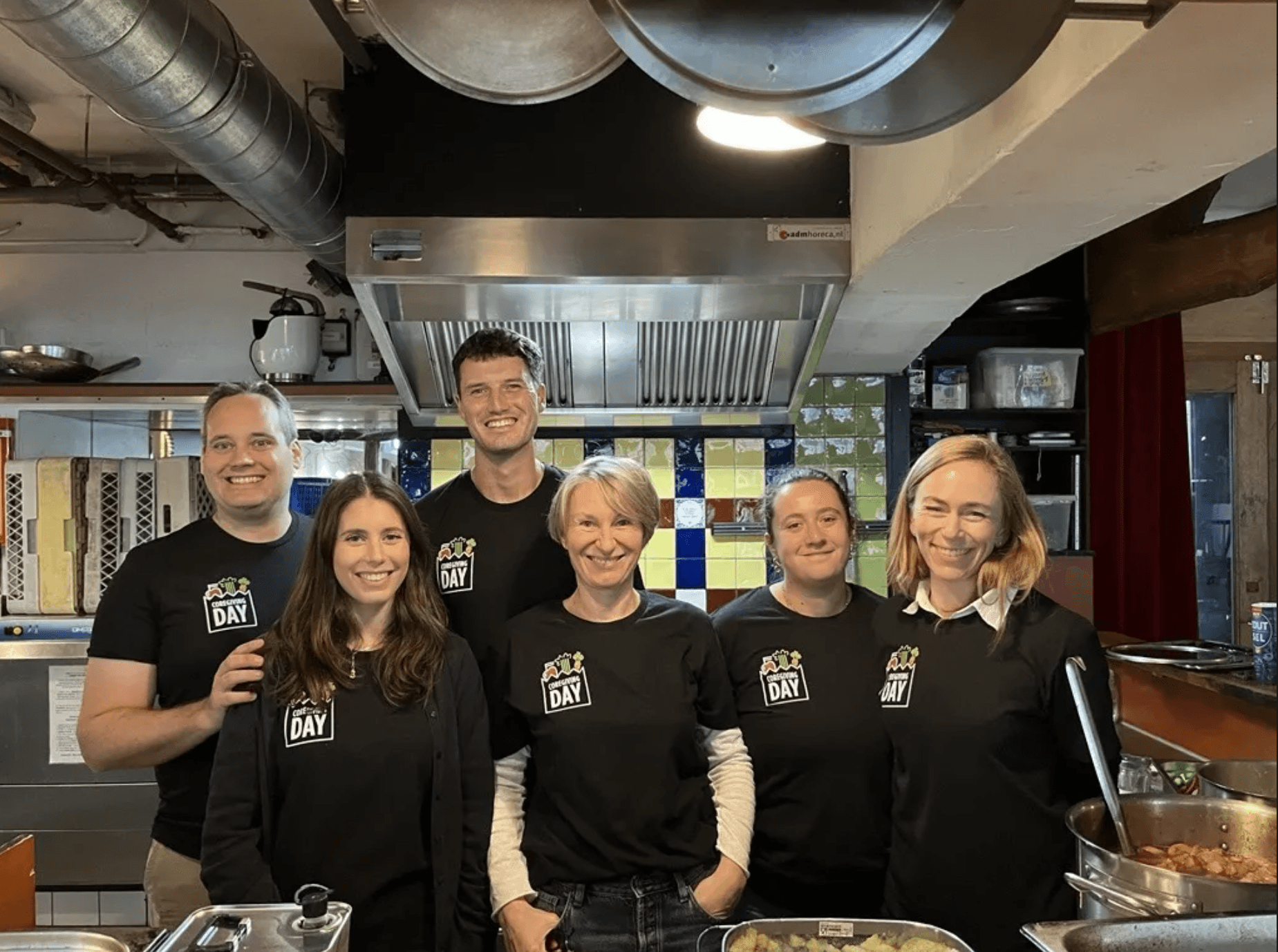 Six people wearing matching black shirts stand together and smile in a commercial kitchen, with food trays visible in the foreground.
