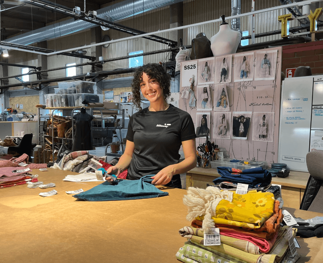 A person stands at a worktable cutting fabric with scissors in a studio, surrounded by fabric stacks and a wall display of garment photos and sketches.