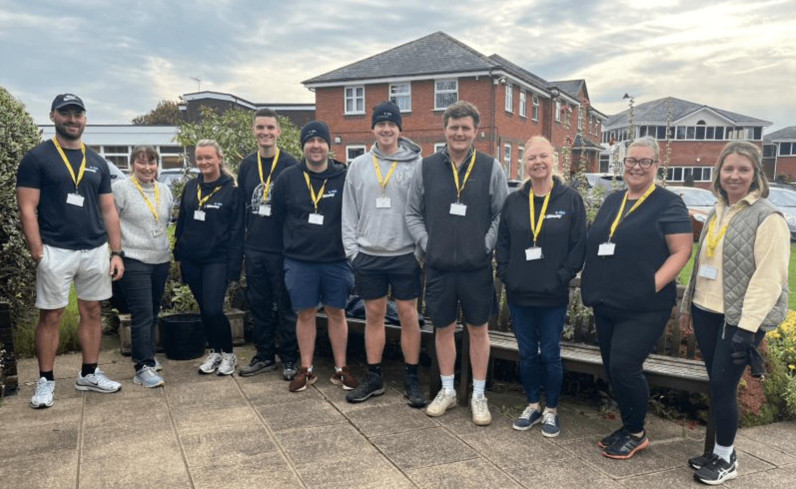 Ten people wearing name badges and yellow lanyards stand in a row outdoors, in front of residential buildings on a cloudy day.