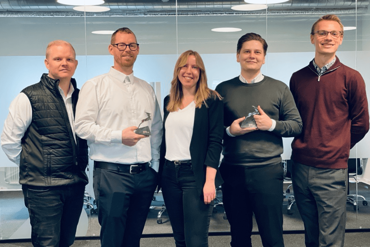 Five people stand indoors in business attire, with two holding awards. They are posed in front of a glass wall in a modern office setting.