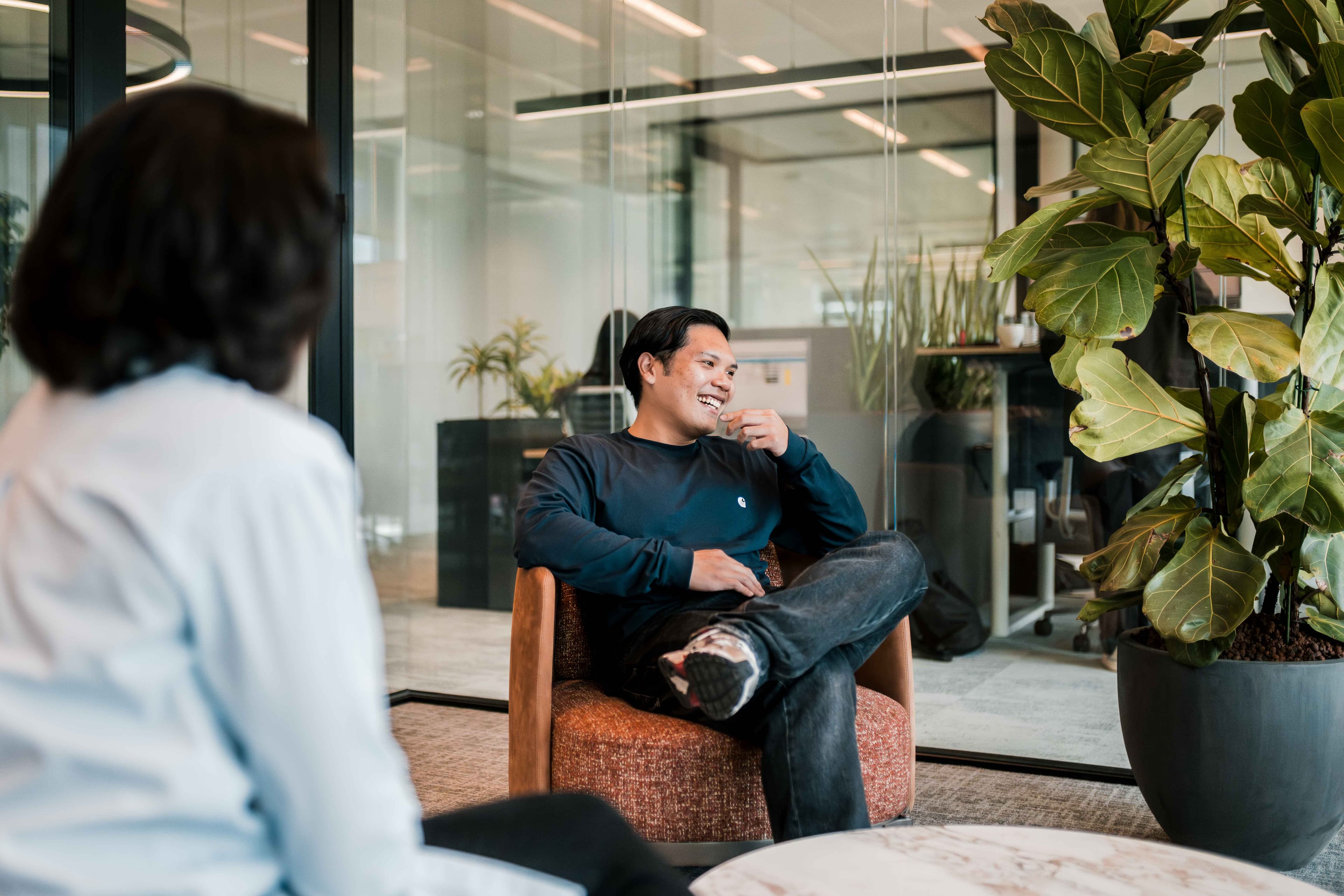 Two people sit and converse in a modern office space with glass walls and large potted plants in the background.