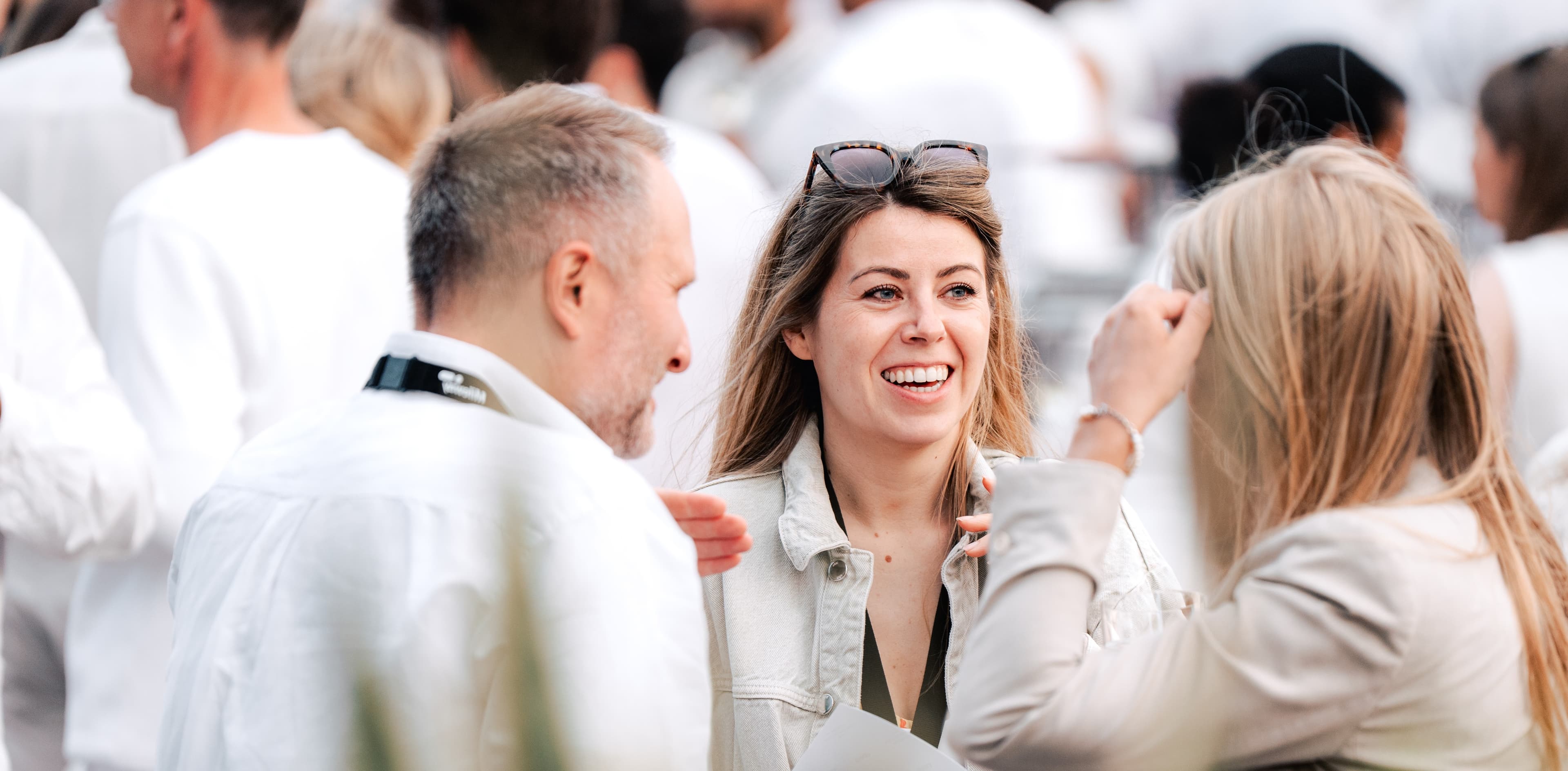 Three people stand outdoors in a crowd, engaged in conversation. The woman in the centre is smiling, and everyone is dressed in light-coloured clothing.