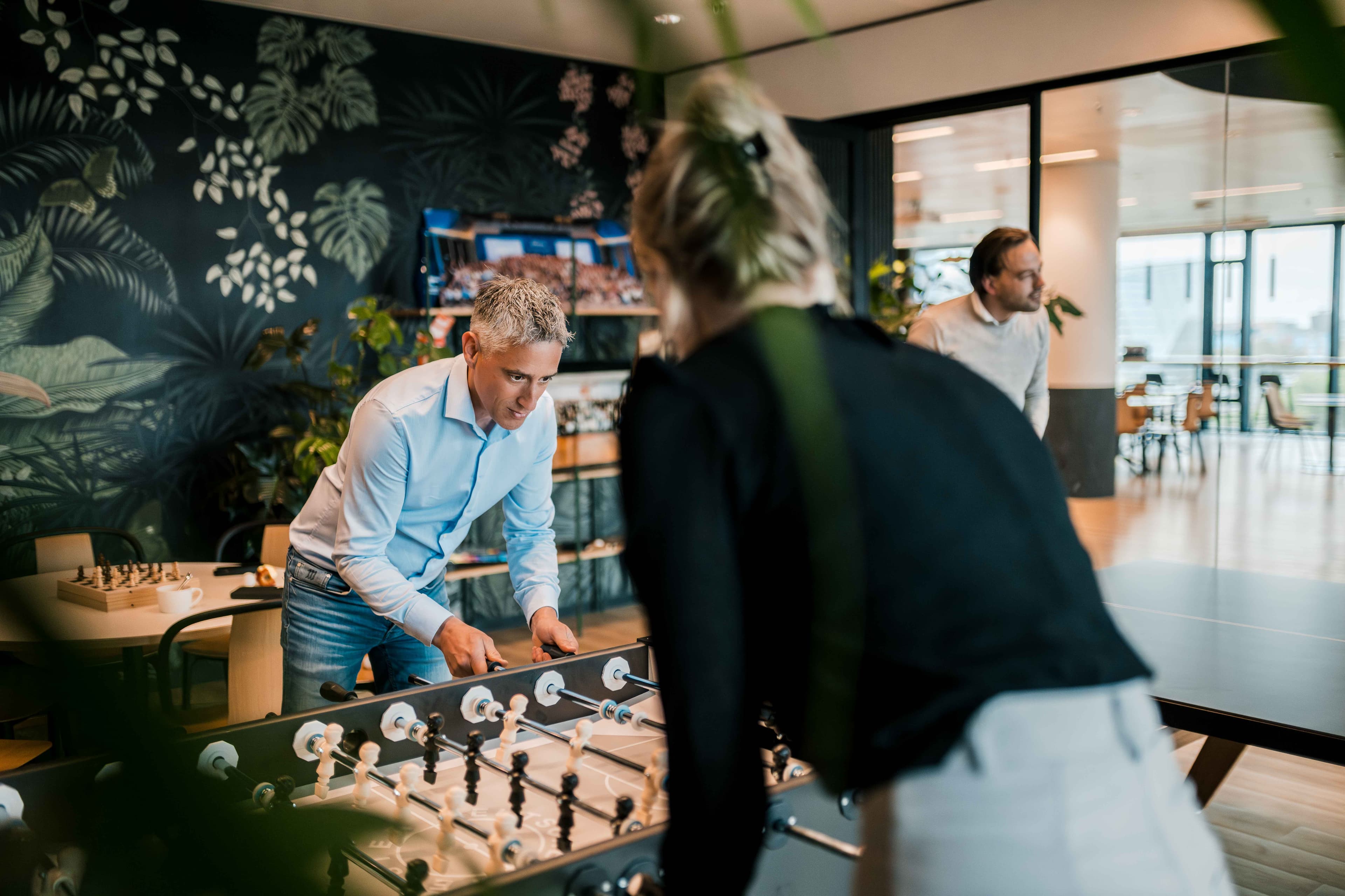 Two people play table football in a modern office lounge while another person stands nearby. The room has large windows, plants, and a dark-patterned feature wall.